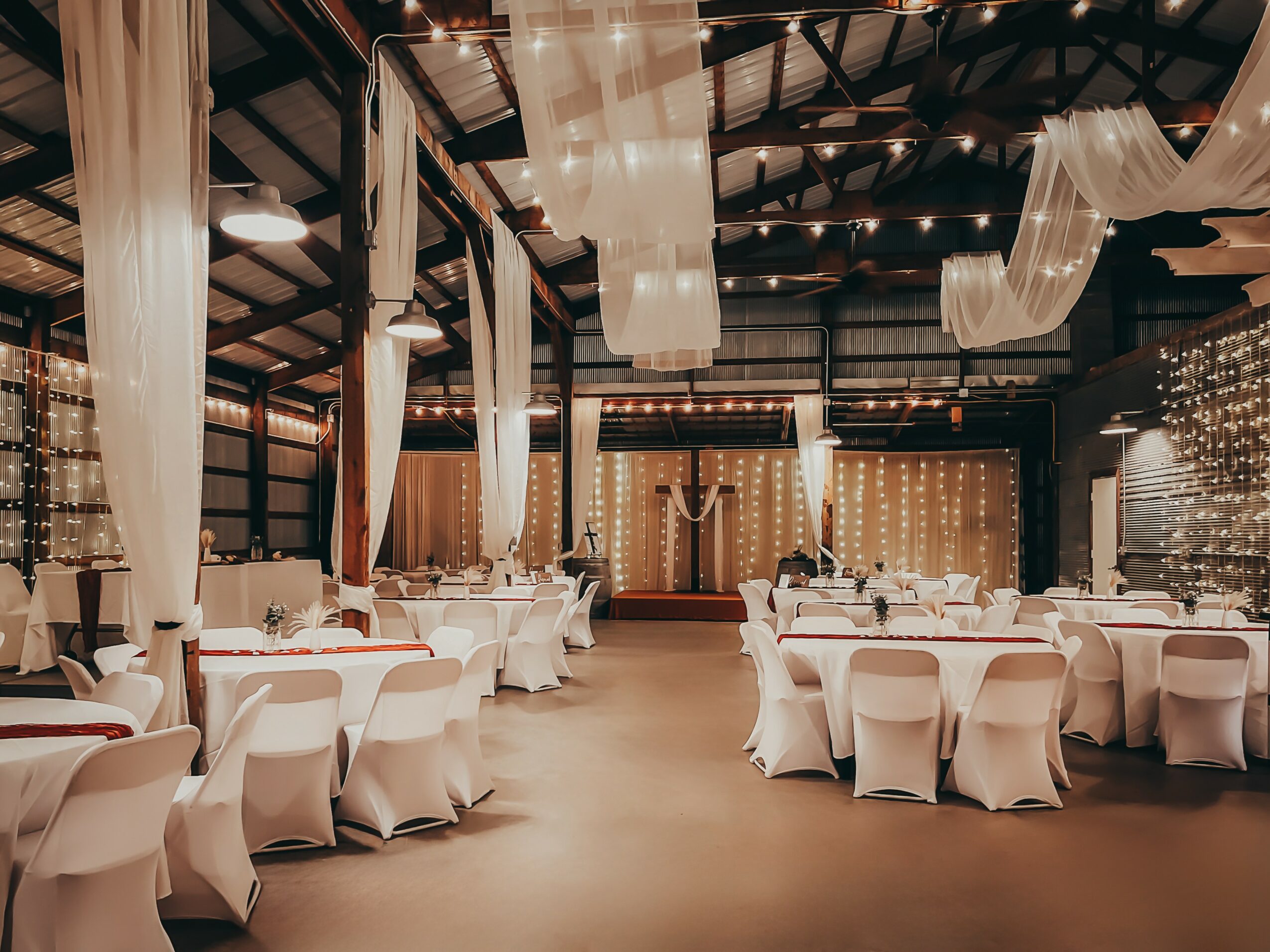Event hall with round tables, white chairs, draped fabric decorations, and a stage with a backdrop, ready for a celebration.