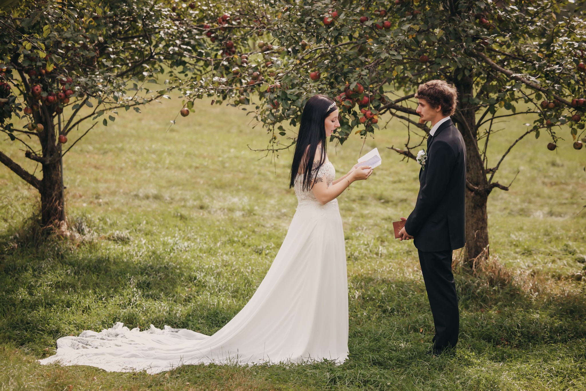 A bride and groom stand outdoors under apple trees, facing each other, with the bride holding a paper, in a grassy area.