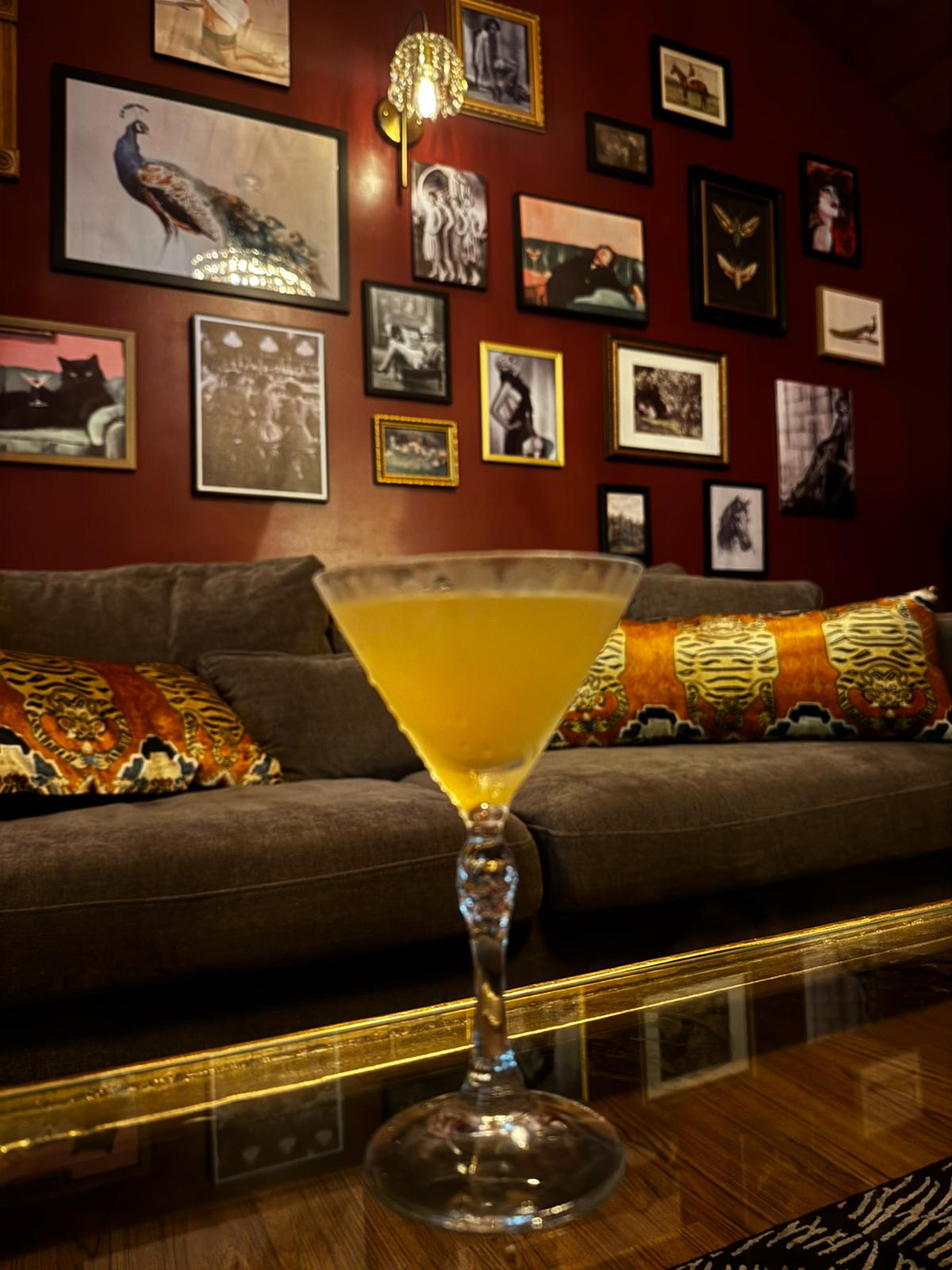 A yellow cocktail in a martini glass on a table, with a sofa and a gallery wall in the background.