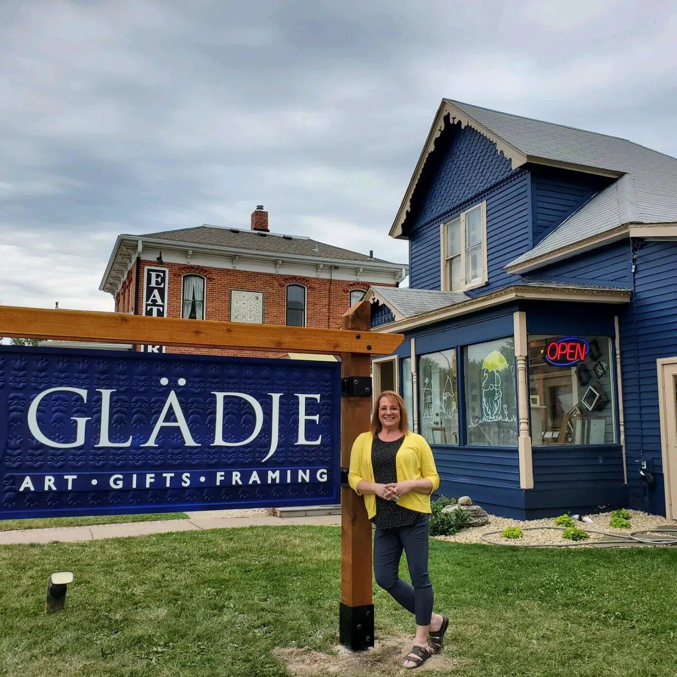 Woman standing outside a blue art store with a sign reading GLÄDJE, holding a book, in front of a building with an open sign.