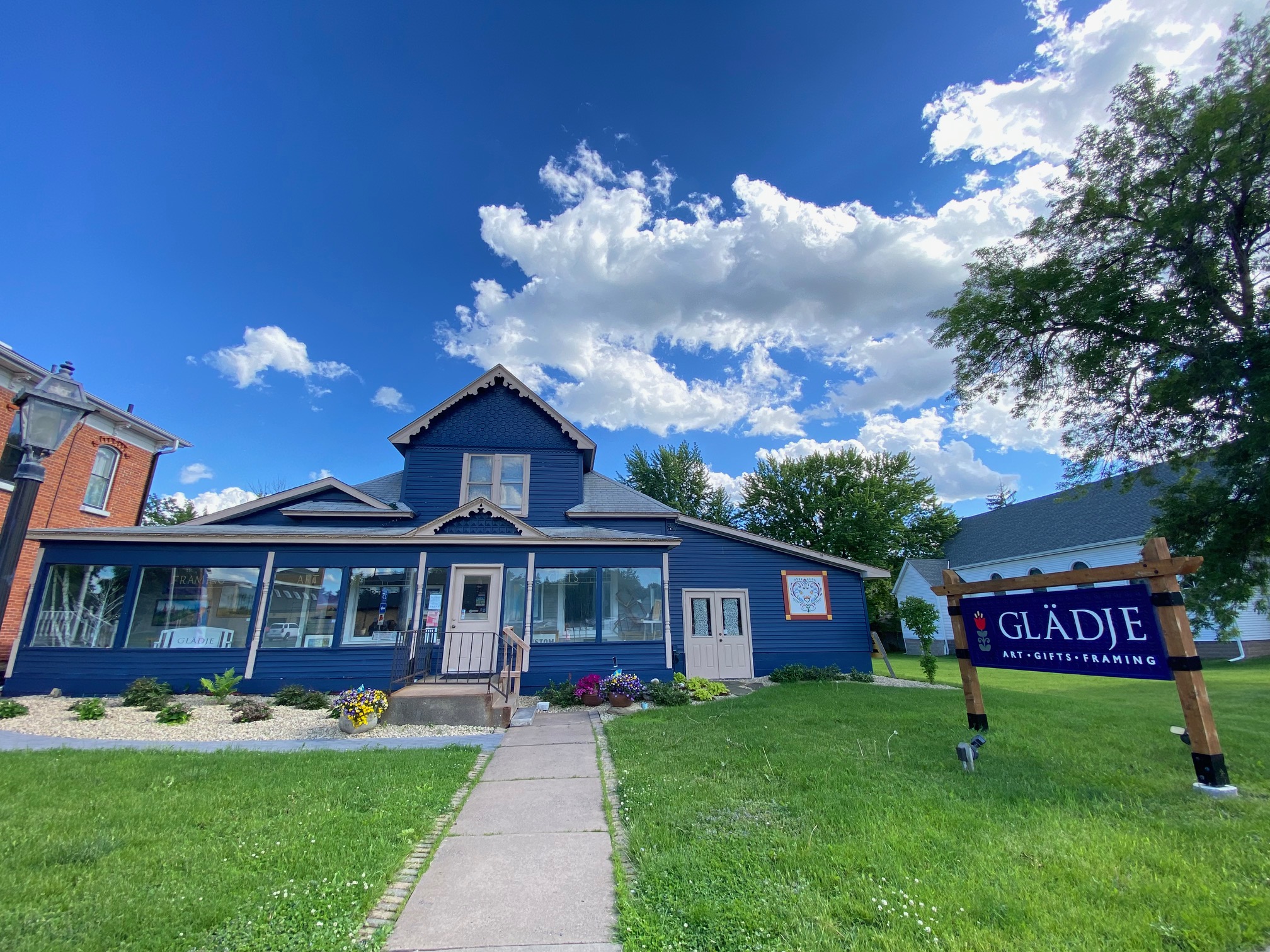 Blue house with a porch, surrounded by green lawn and trees, under a partly cloudy sky.