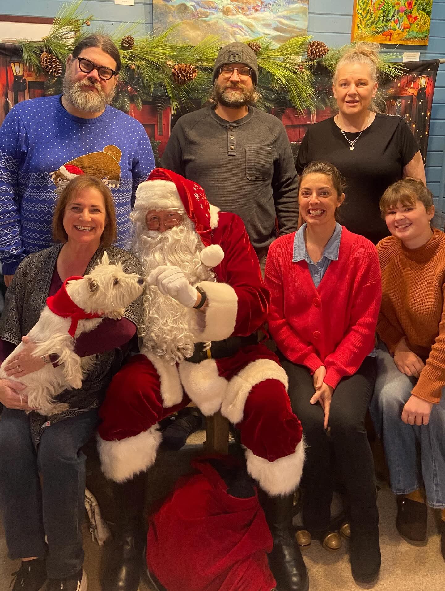 Group of seven people, including a person dressed as Santa Claus, posing indoors with festive decorations and two dogs.