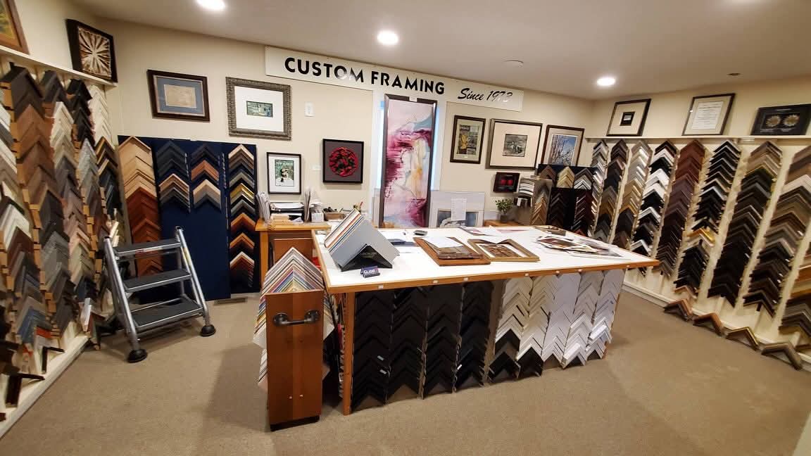 Interior of a custom framing shop with display racks of framing samples, a central table with framing materials, and framed artwork on walls.