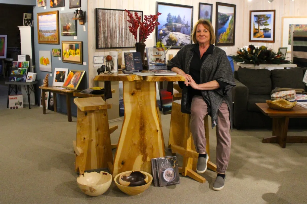 Woman standing next to a wooden table with three matching stools in an art gallery, surrounded by framed paintings and decor.