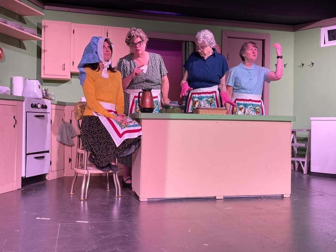Four women in a kitchen, one sitting on a chair, three standing, with pastel-colored cabinets and appliances.