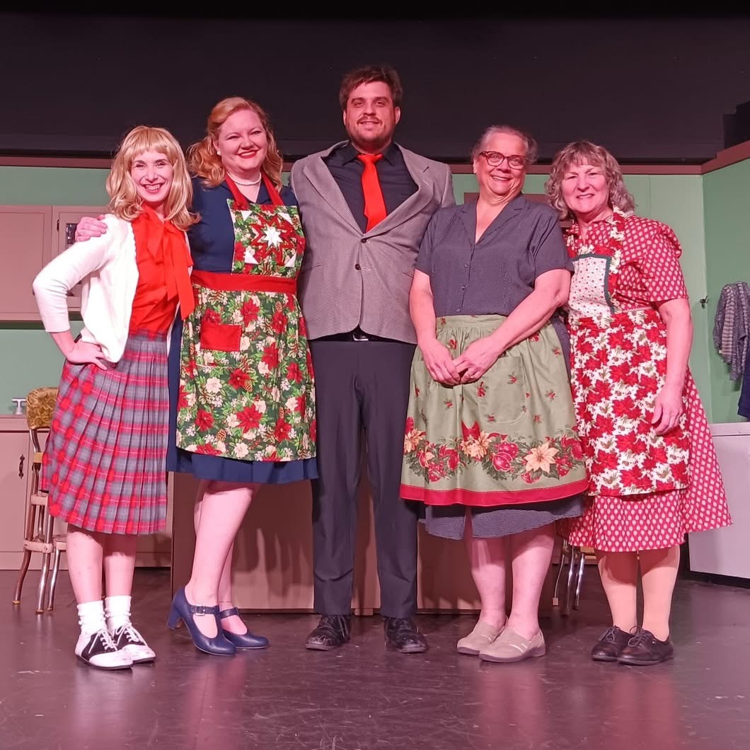 Five people standing on stage, smiling, with a plain background, dressed in colorful vintage-style clothing.