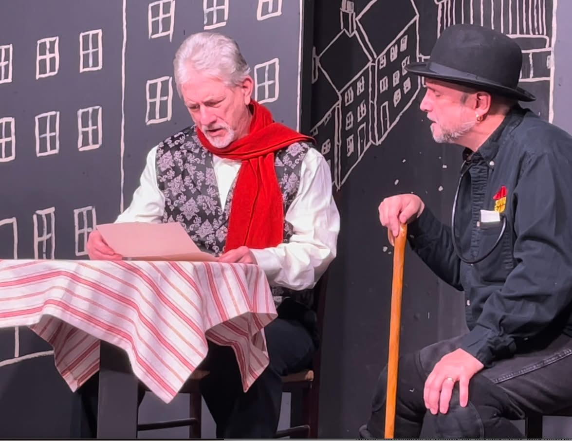 Two men sitting at a table with a striped tablecloth, one reading a book, the other holding a cane, in front of a cityscape backdrop.