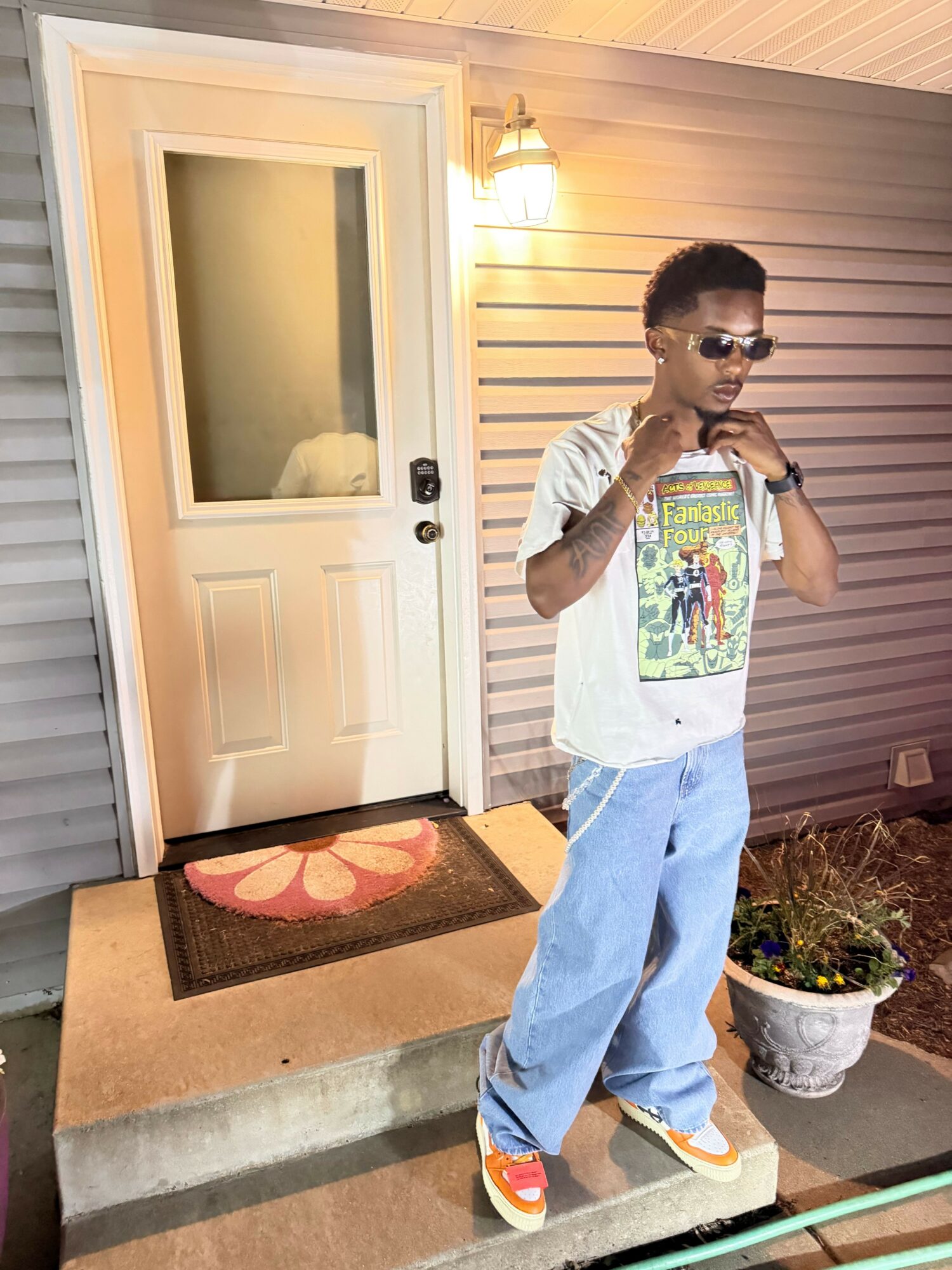 Person standing outside near a door, wearing sunglasses, a graphic t-shirt, light blue jeans, and sneakers, holding a phone.