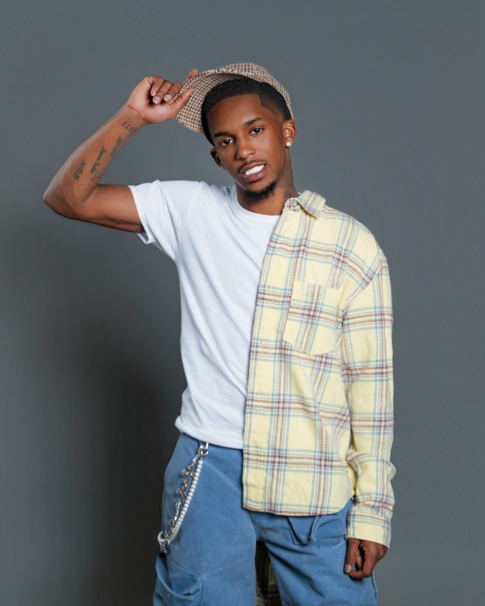 Young man wearing a white t-shirt, blue pants, a plaid shirt draped over shoulder, and a hat, standing against a gray background.