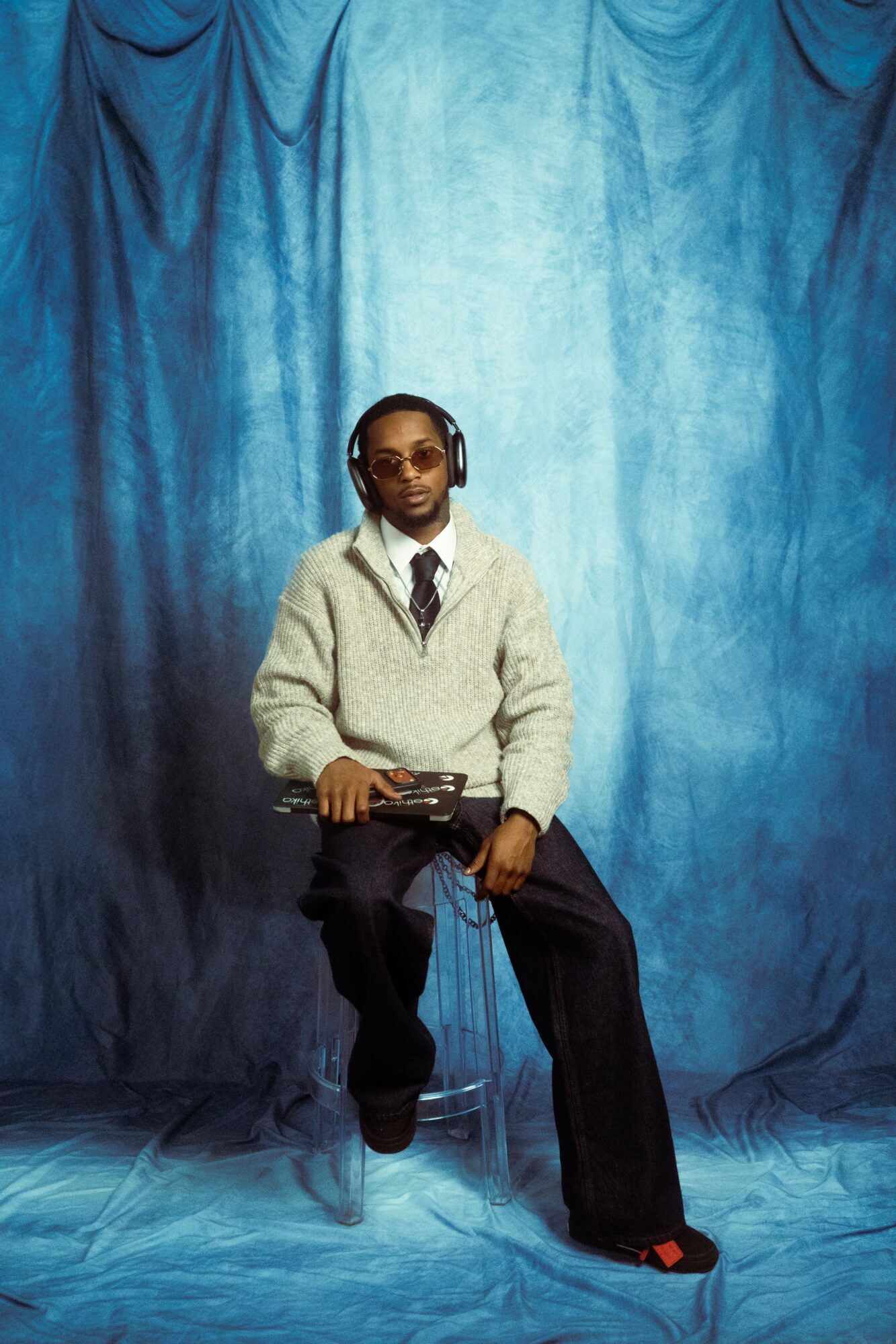 Person sitting on a chair with headphones, wearing glasses, a sweater, and a tie, against a blue curtain backdrop.