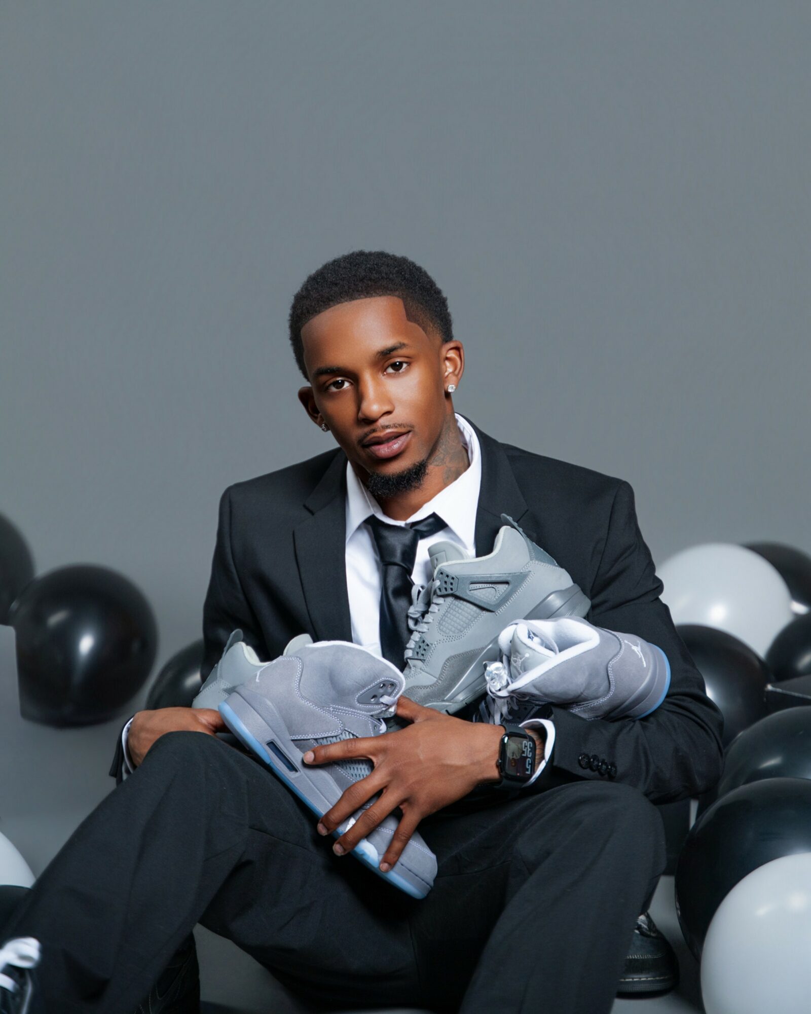 Young man in a suit holding sneakers, sitting among black and white balloons, against a gray background.