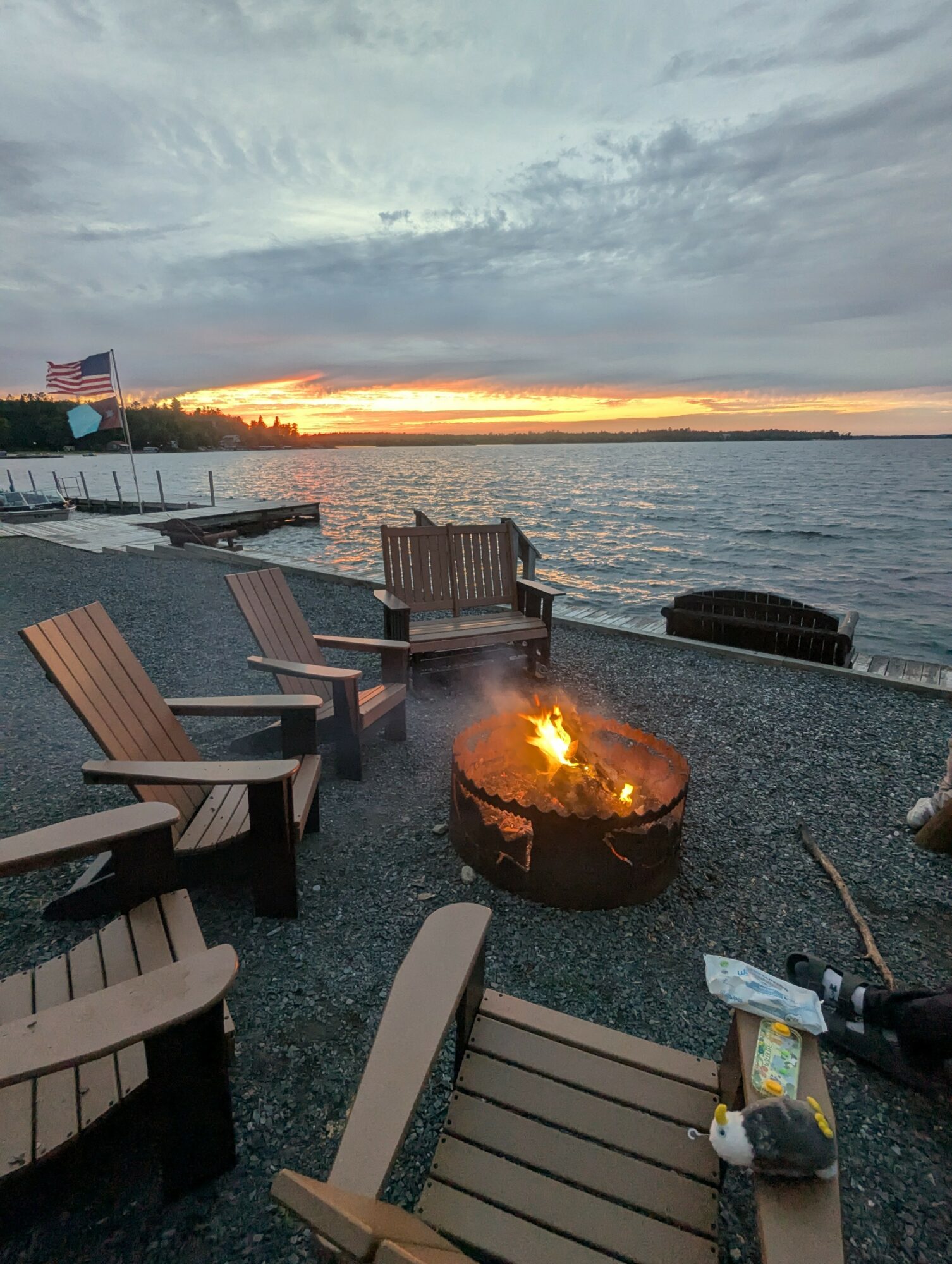 Seaside scene with Adirondack chairs around a fire pit, sunset over water, cloudy sky, American flag in background.