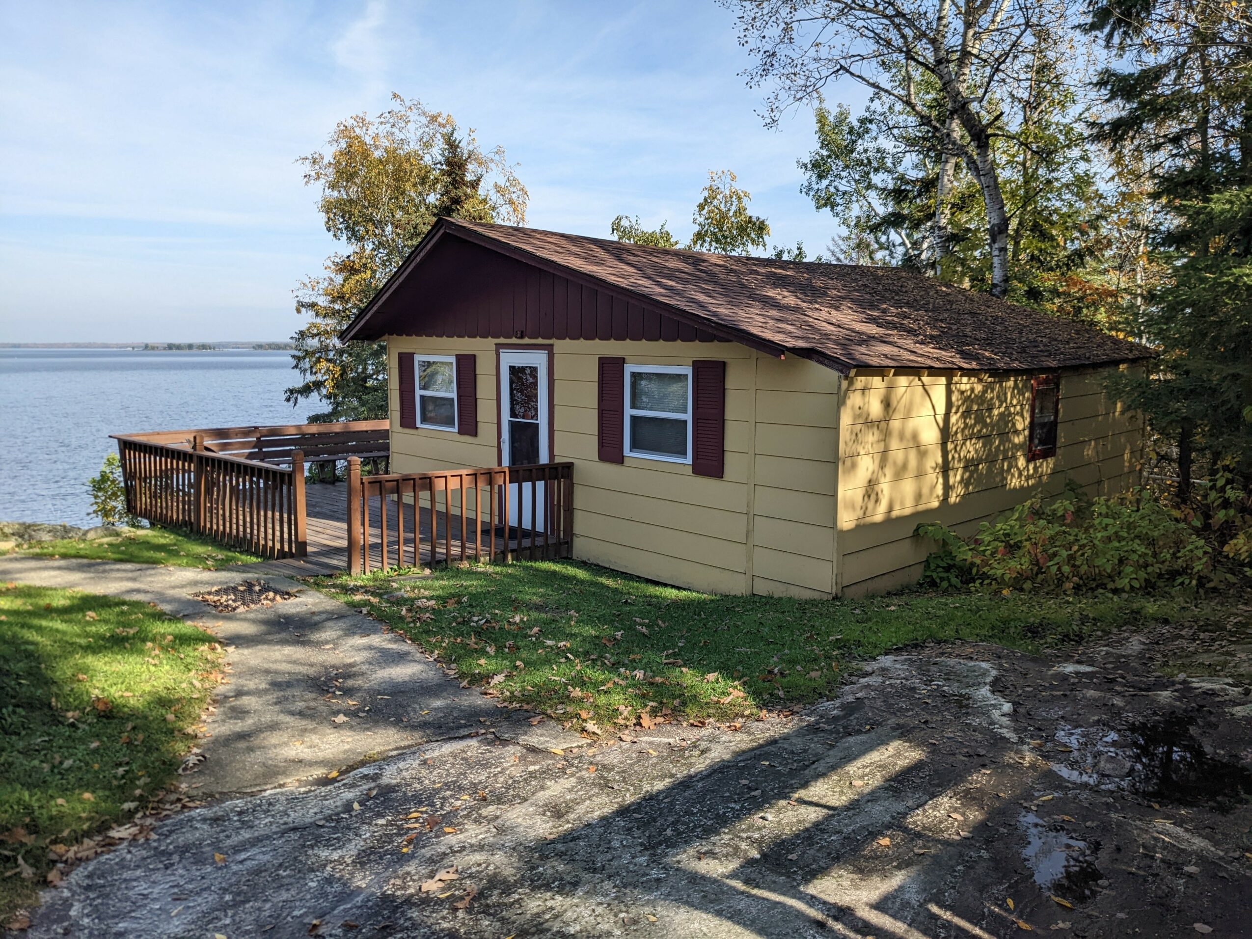Small yellow house with brown roof and shutters near water, surrounded by trees, with a wooden deck and pathway.