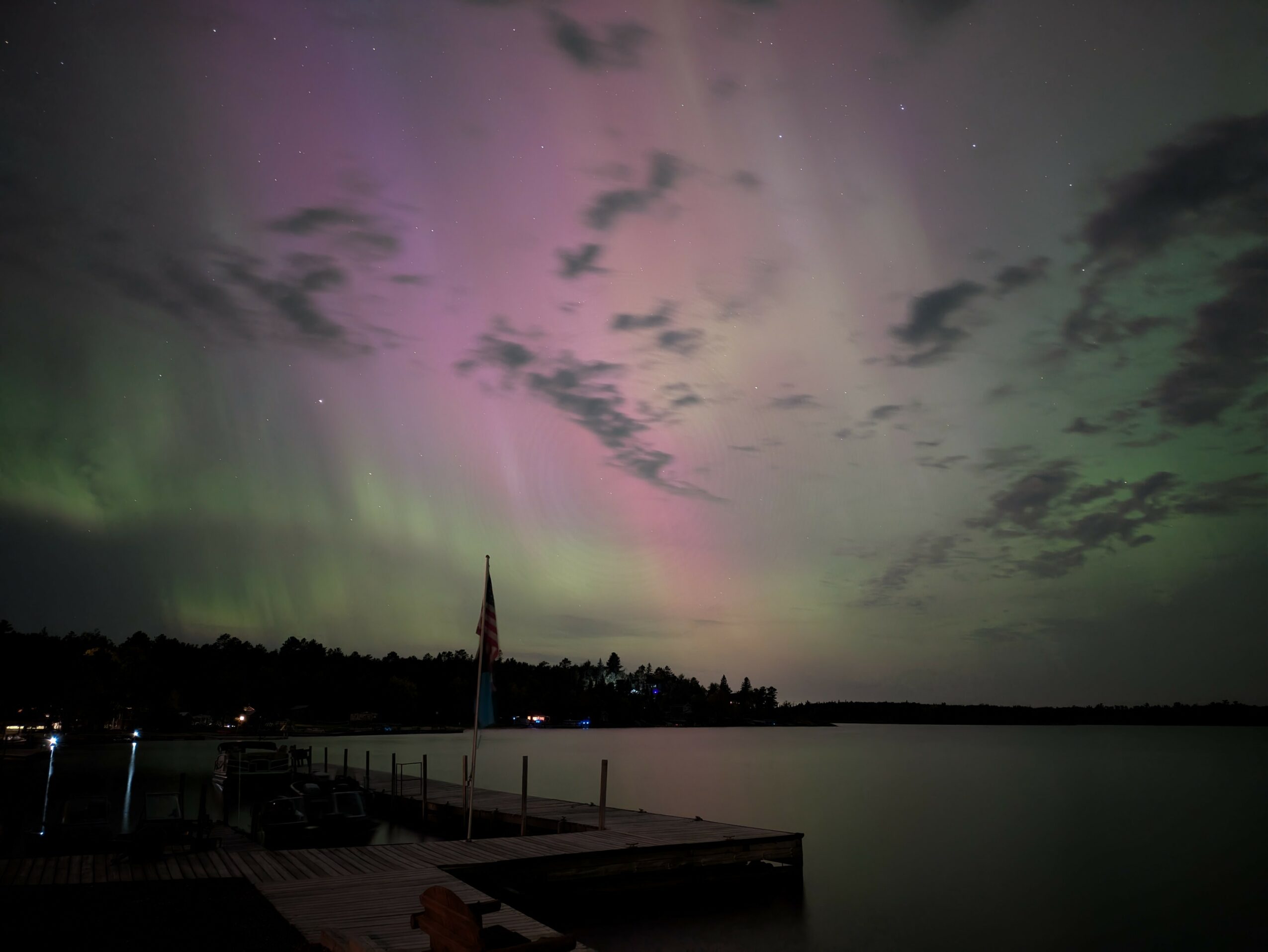 Night sky with colorful aurora borealis over a body of water, with a dock and flag in the foreground.