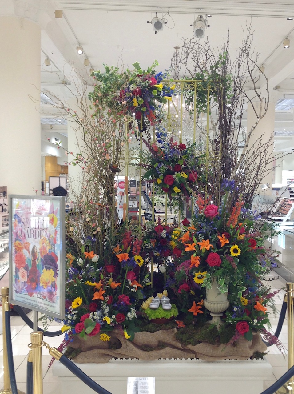Flower arrangement with colorful flowers and branches in a white vase inside a shopping mall, with a display board nearby.