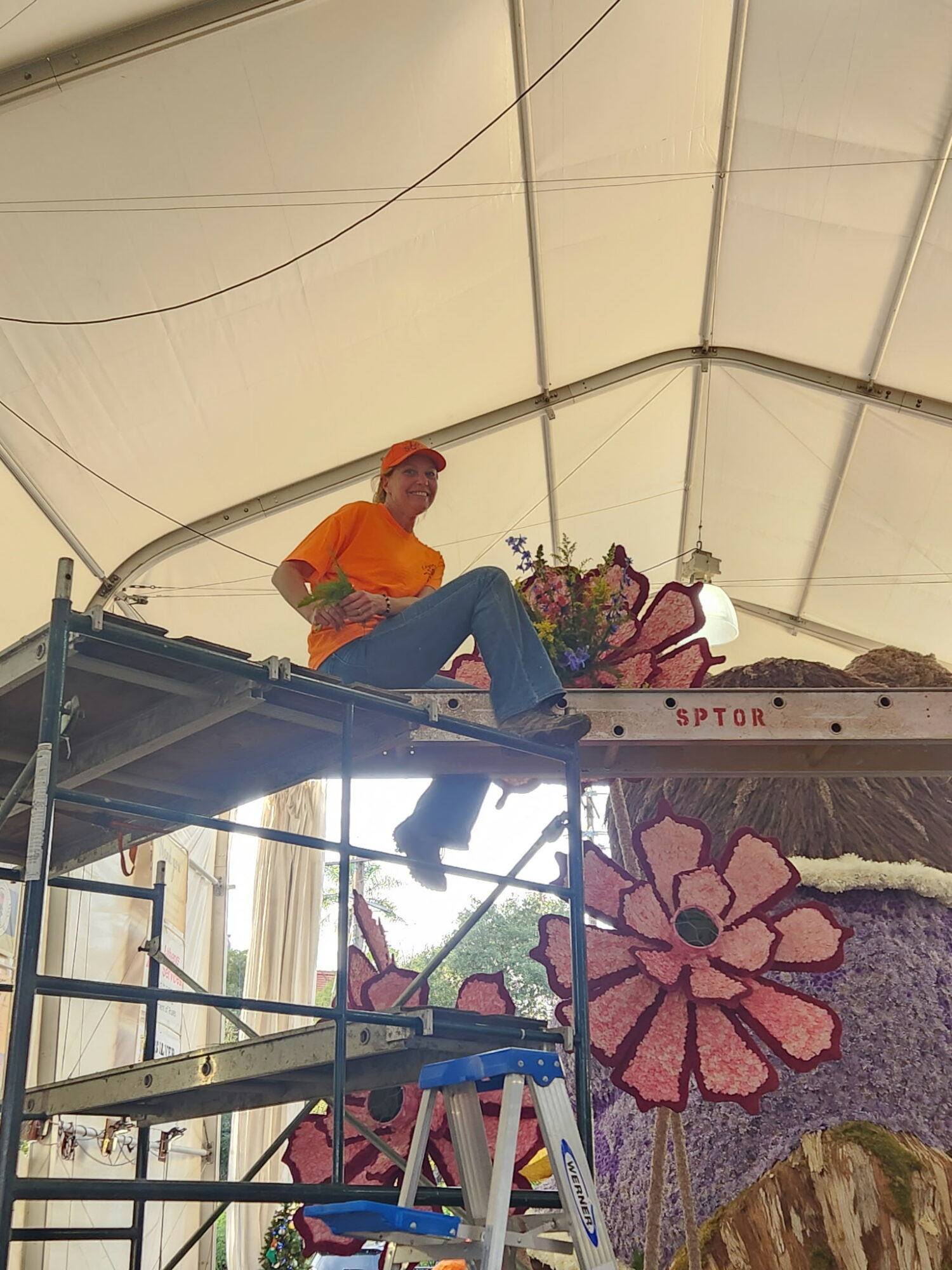 Person sitting on a platform inside a tent, surrounded by colorful floral decorations, wearing an orange shirt and blue jeans.