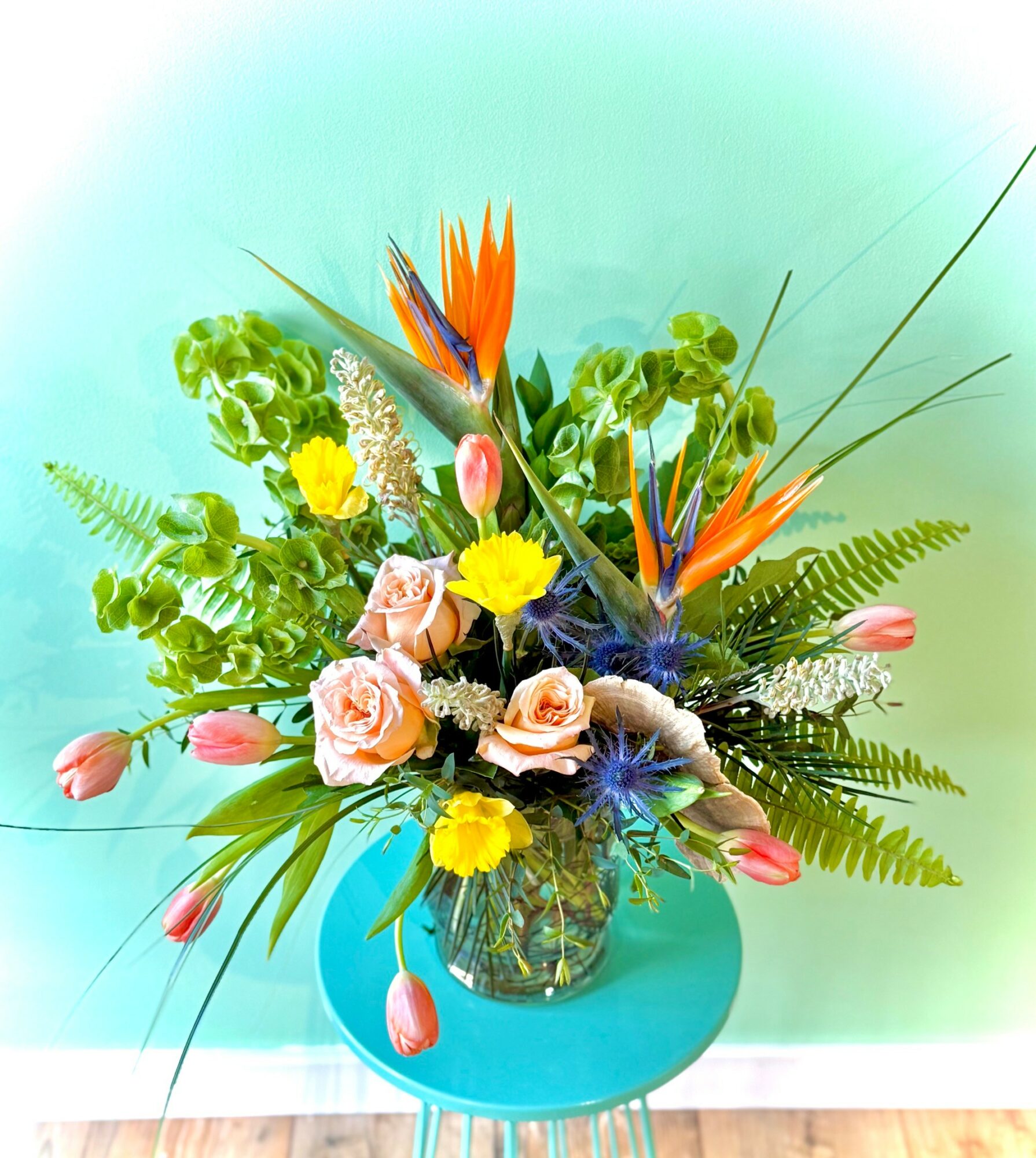Colorful flower arrangement with roses, tulips, and exotic flowers in a blue vase on a round table, against a green wall.
