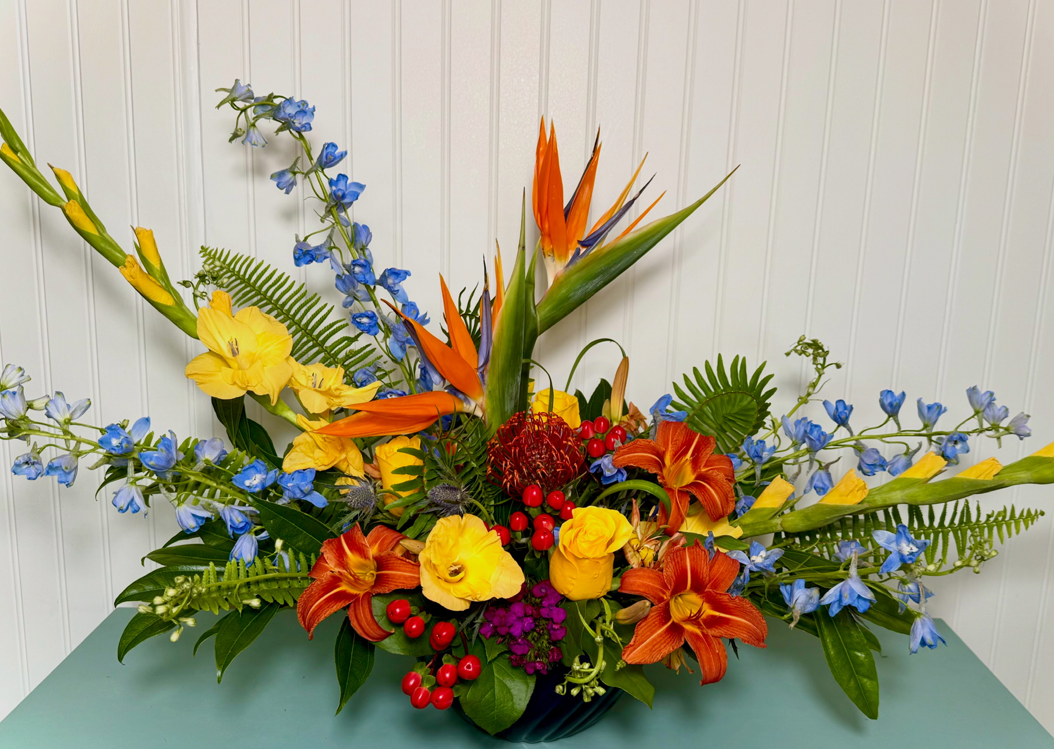 Colorful flower arrangement with orange, yellow, red, purple, and blue flowers and green foliage, placed against a white background.