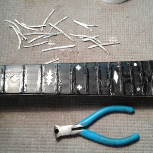 Broken glass pieces above a guitar fretboard and blue-handled pliers on a textured surface.