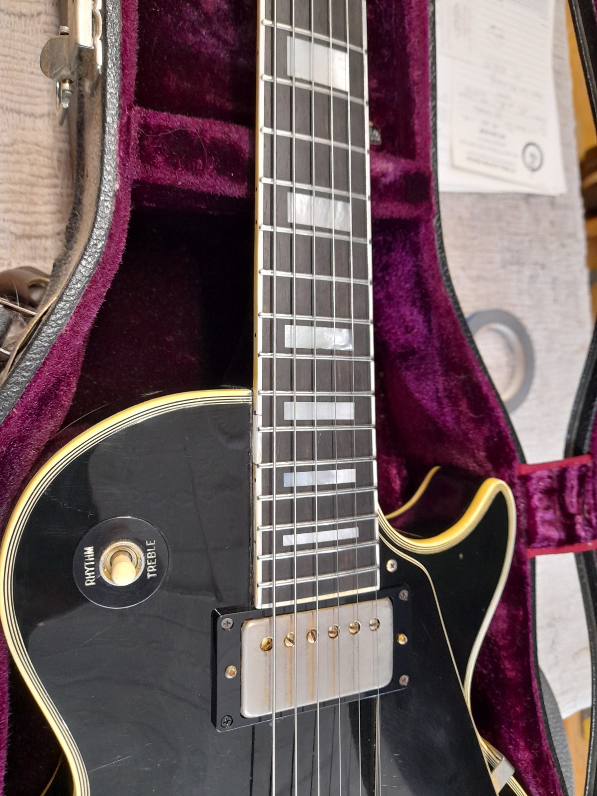 Black electric guitar with a gold bridge and pickups, inside a purple-lined case.