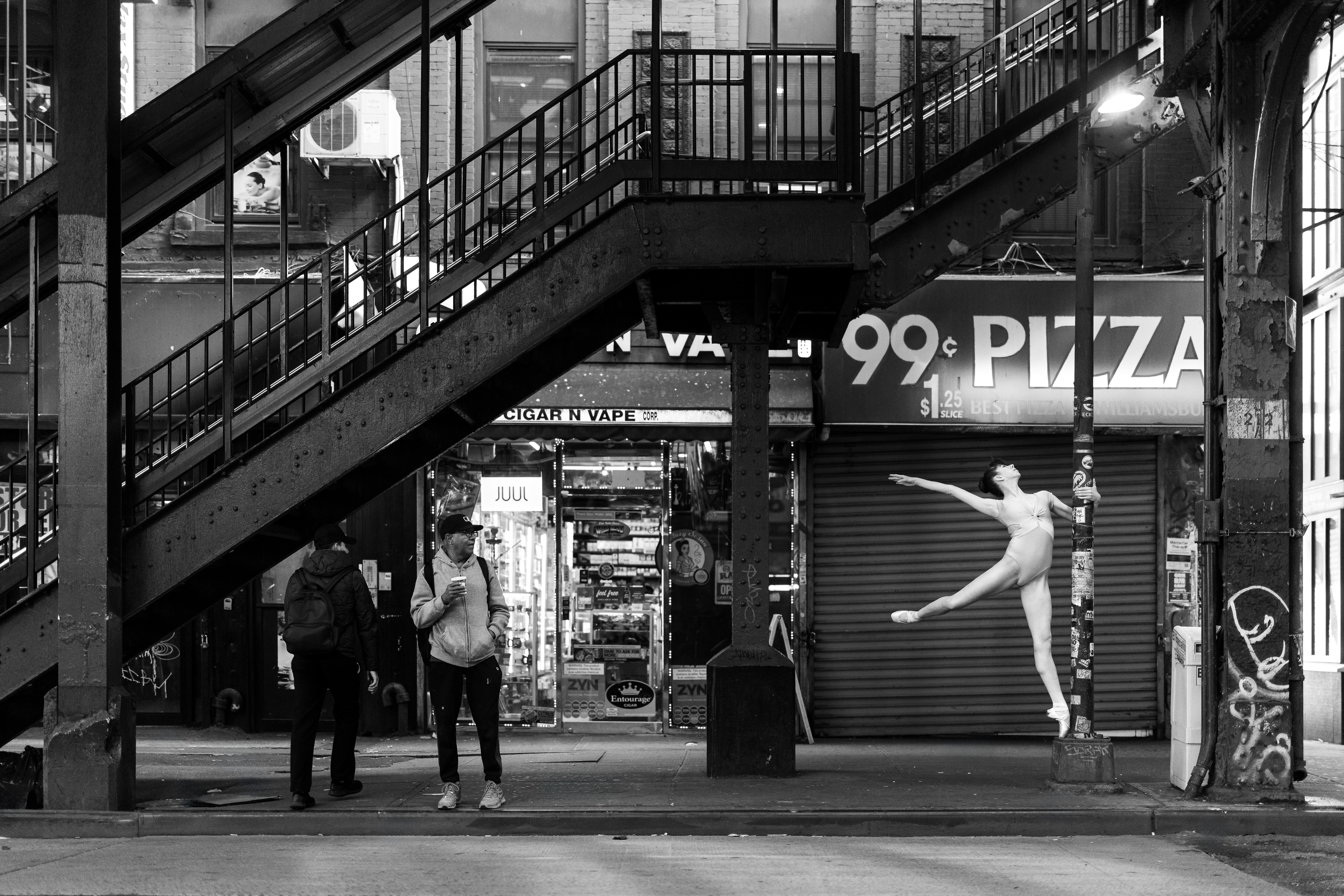 Street scene with people walking past a closed pizza shop and a ballet dancer figure on the shutter, staircase overhead.
