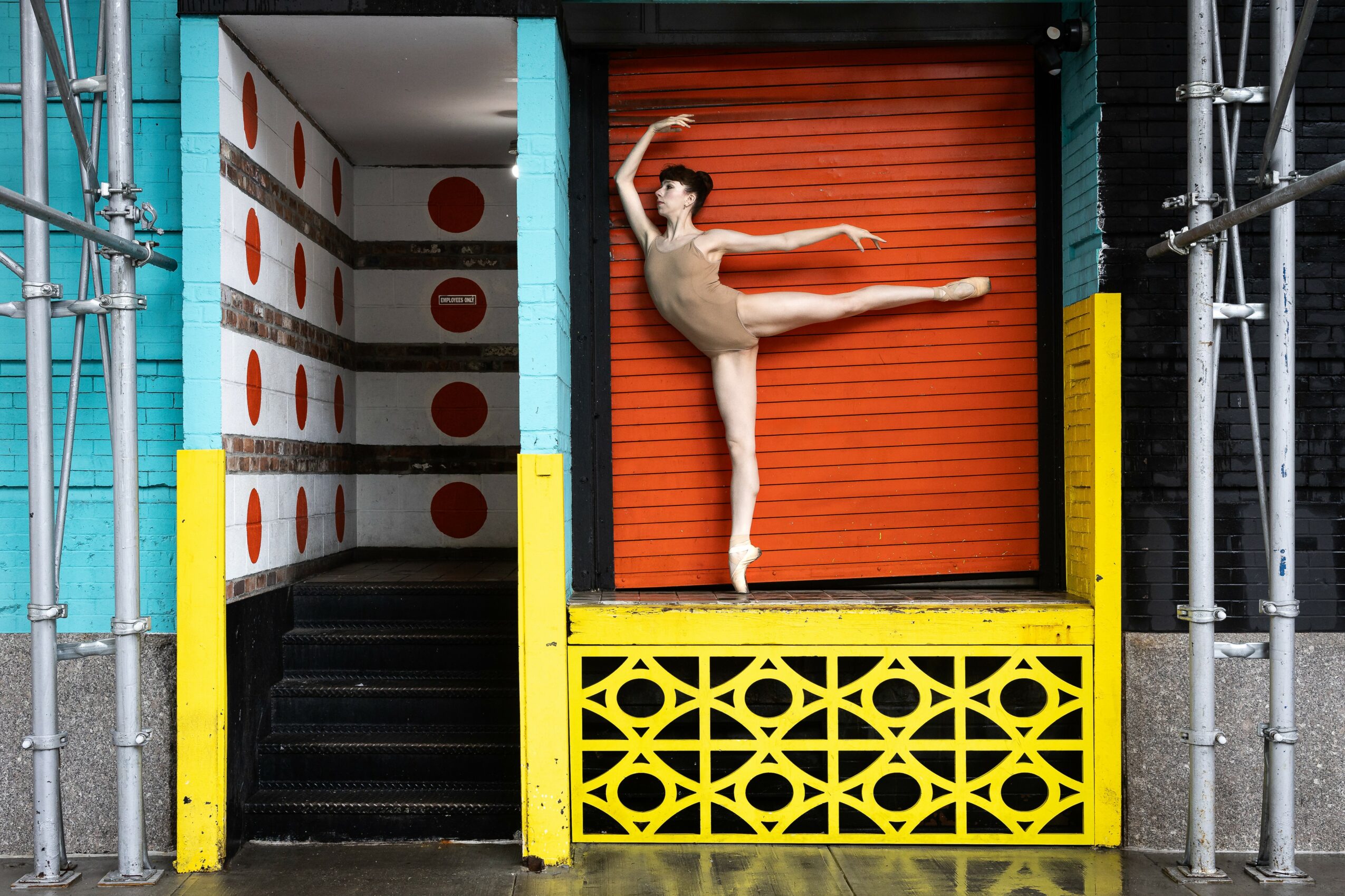 Ballet dancer in a beige costume performs a pose on an orange rolling shutter, with scaffolding and colorful structures around.