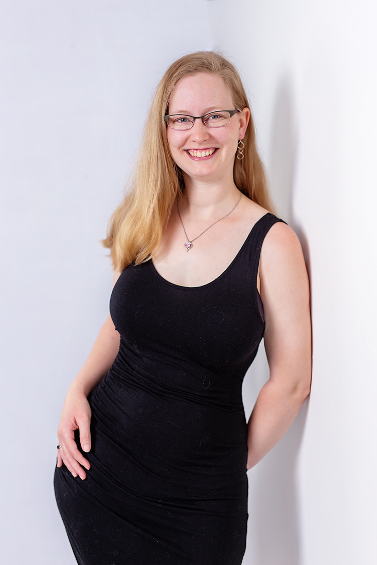 Smiling woman with glasses and long blonde hair wearing a black dress, standing against a white wall.