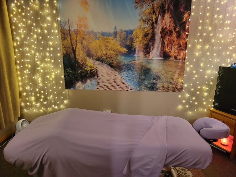 A bed with a light purple sheet in a room with a nature scene tapestry and string lights on the wall.