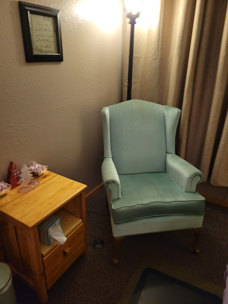 Light blue armchair with high back and armrests next to a wooden side table, in a room with beige walls and curtains.