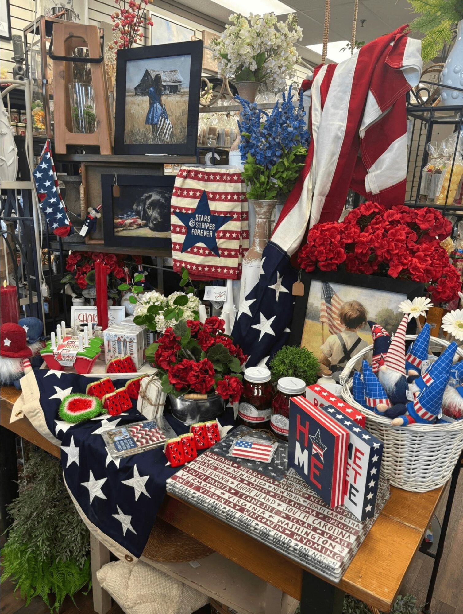 Table with patriotic decorations including flags, flowers, and framed photos, featuring red, white, and blue items and an American flag-themed pillow.