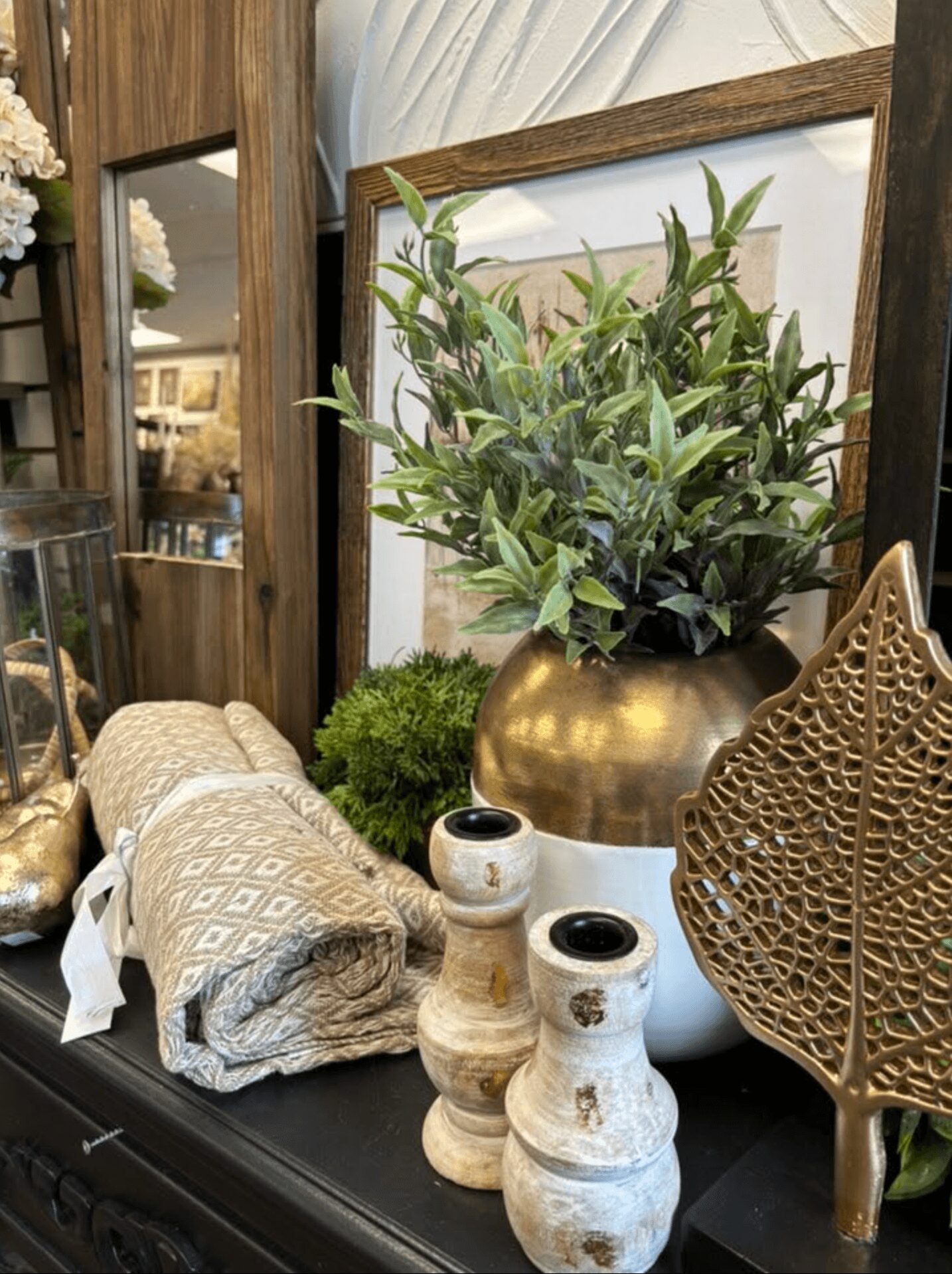 Decorative arrangement with a large gold vase, green foliage, textured vases, and a woven decorative object on a dark surface.