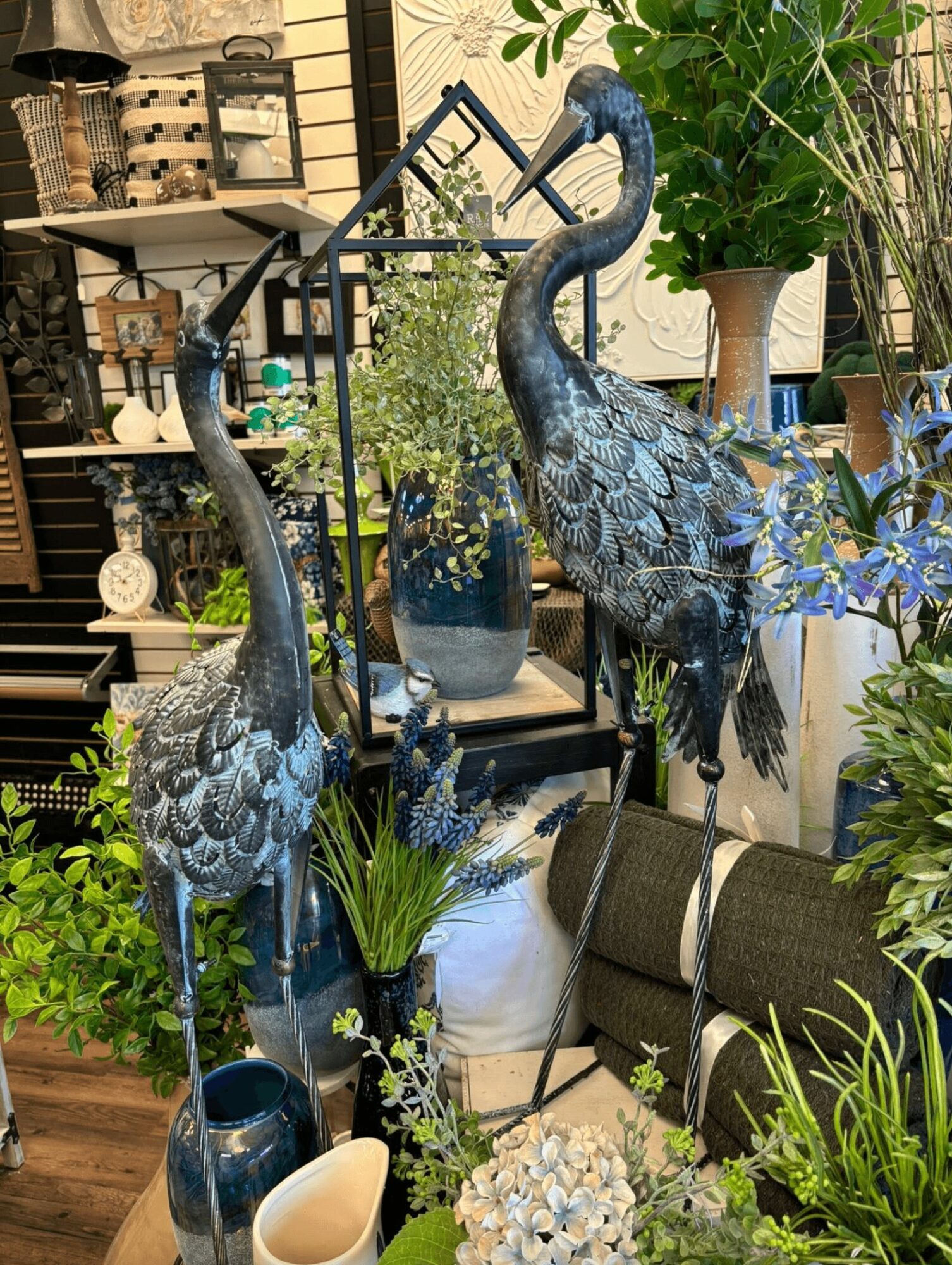 Two decorative blue heron statues surrounded by potted plants and garden decor in an indoor setting.