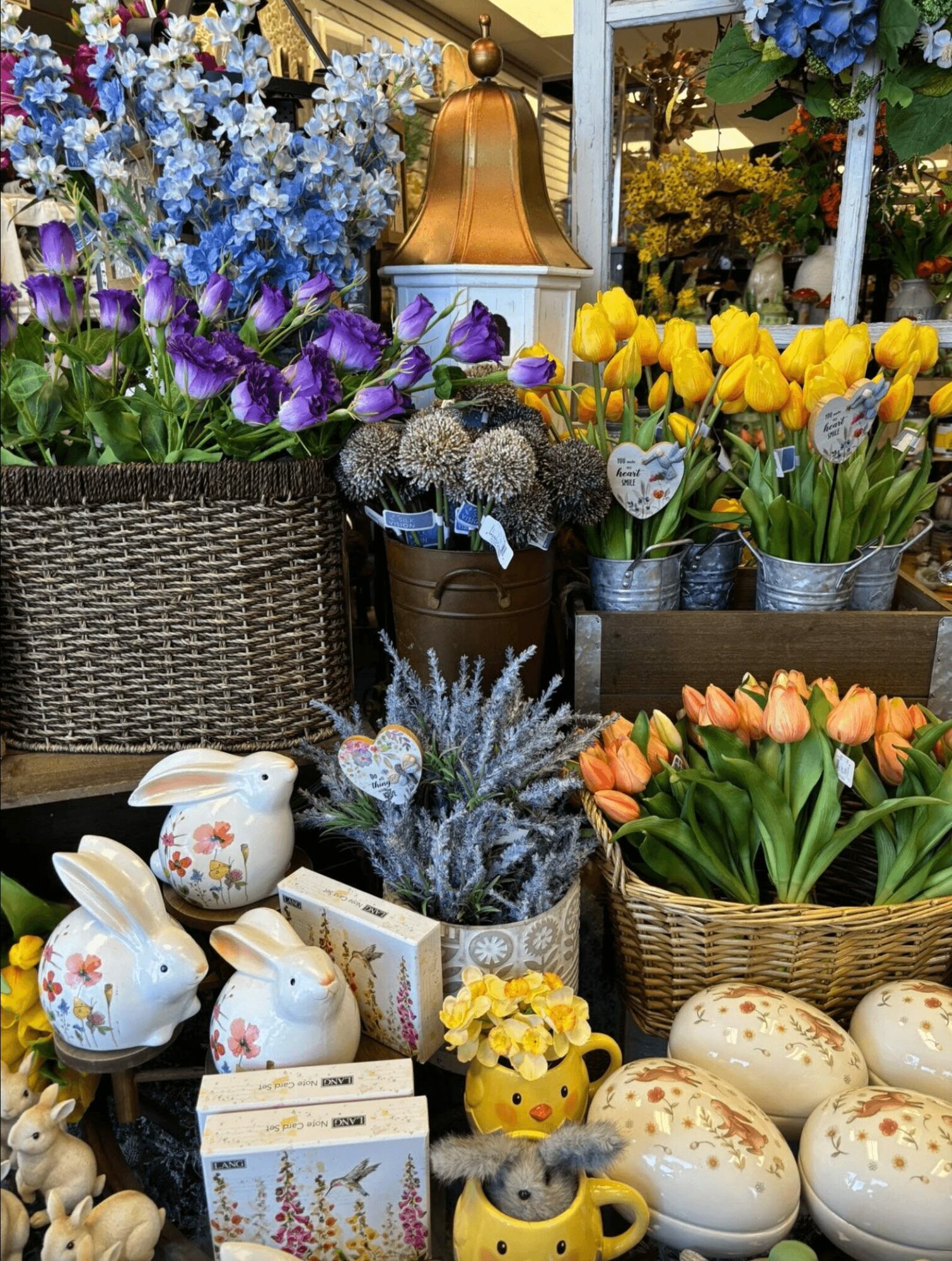 Colorful flowers including purple, yellow, orange, and blue arranged in baskets and pots with decorative eggs and bunny figurines.