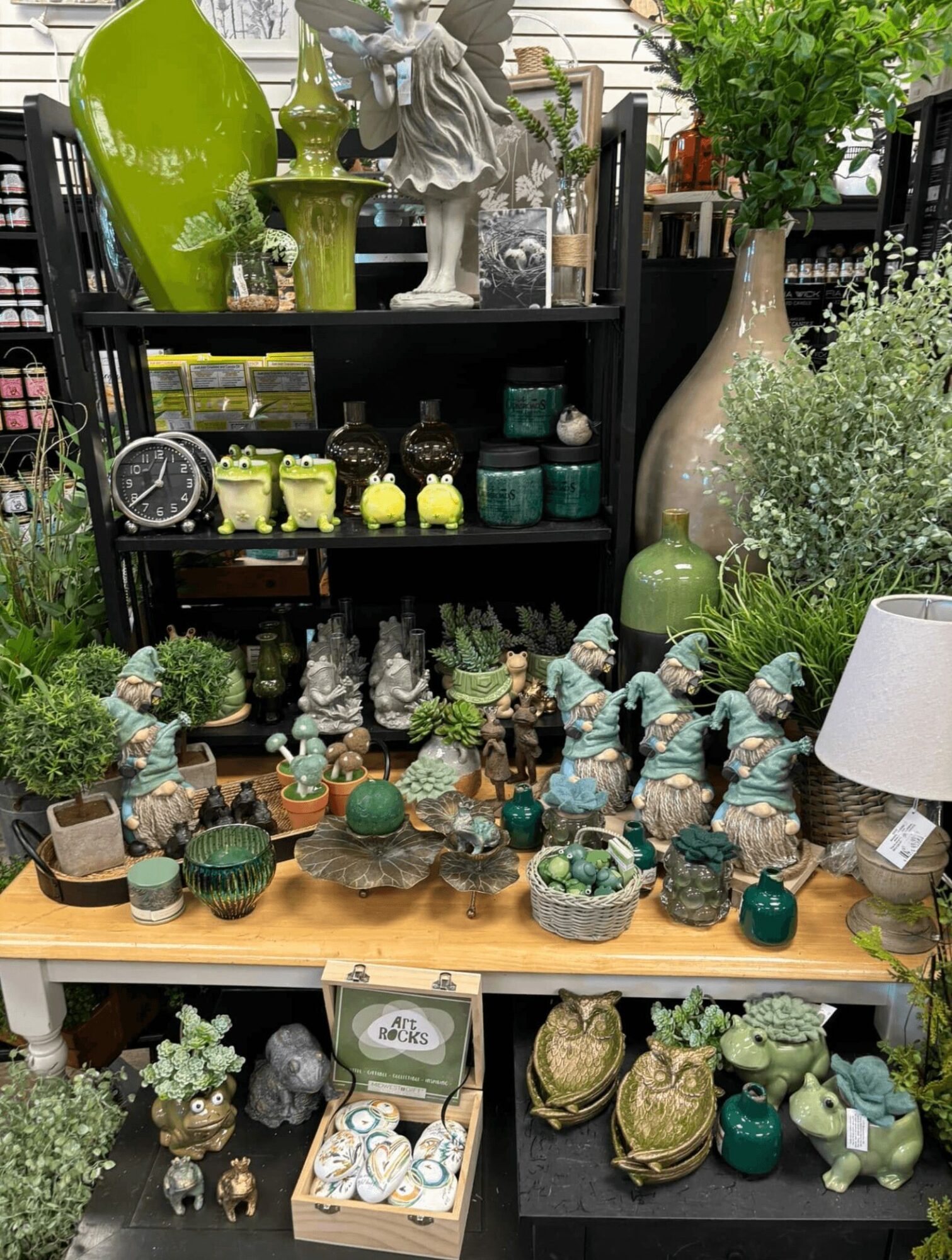 Display of various decorative items including vases, figurines, plants, and ceramics on shelves and a table, with greenery in the background.