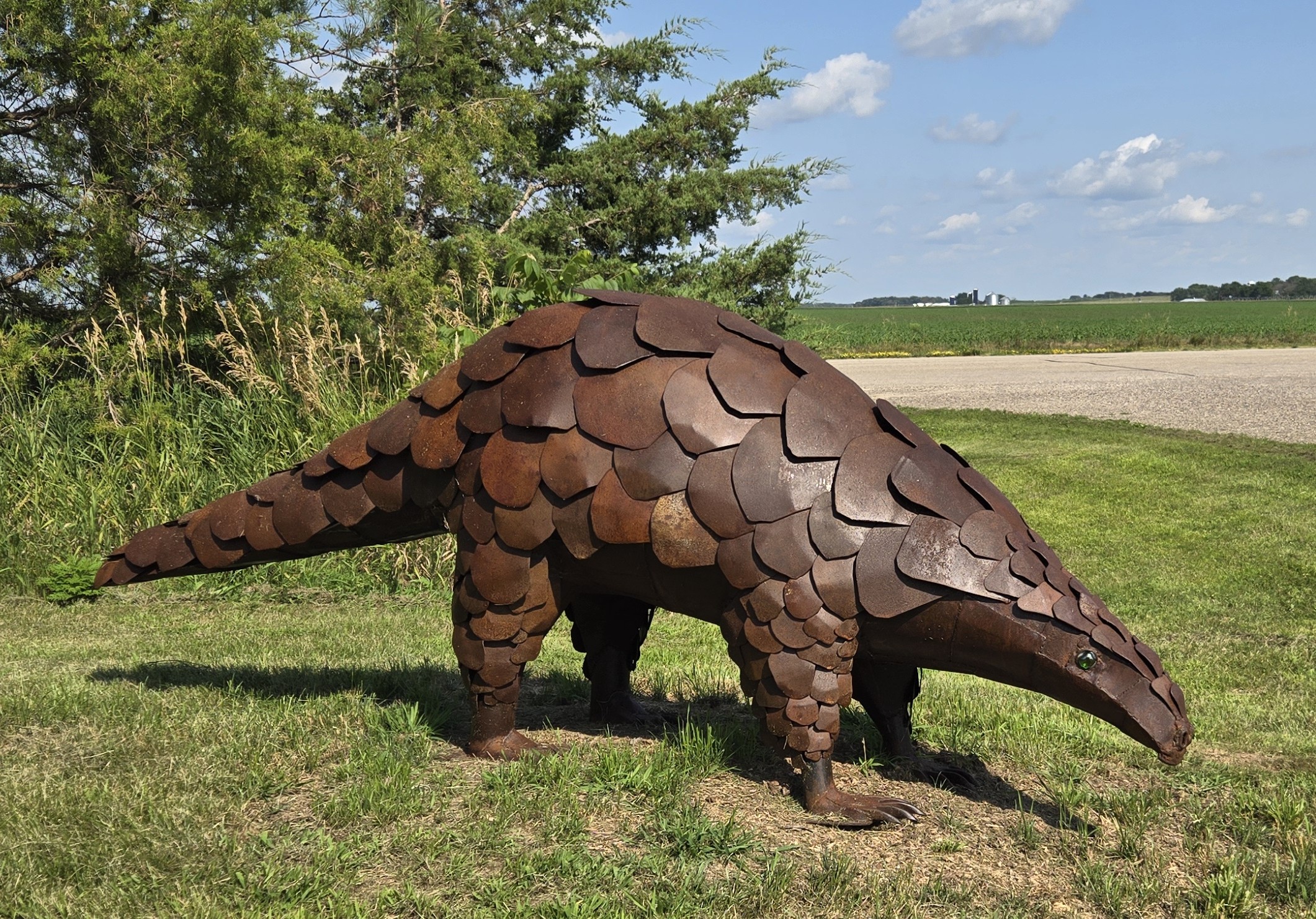 A sculpture of an armadillo made from overlapping metal plates, outdoors on grass with trees and sky in background.