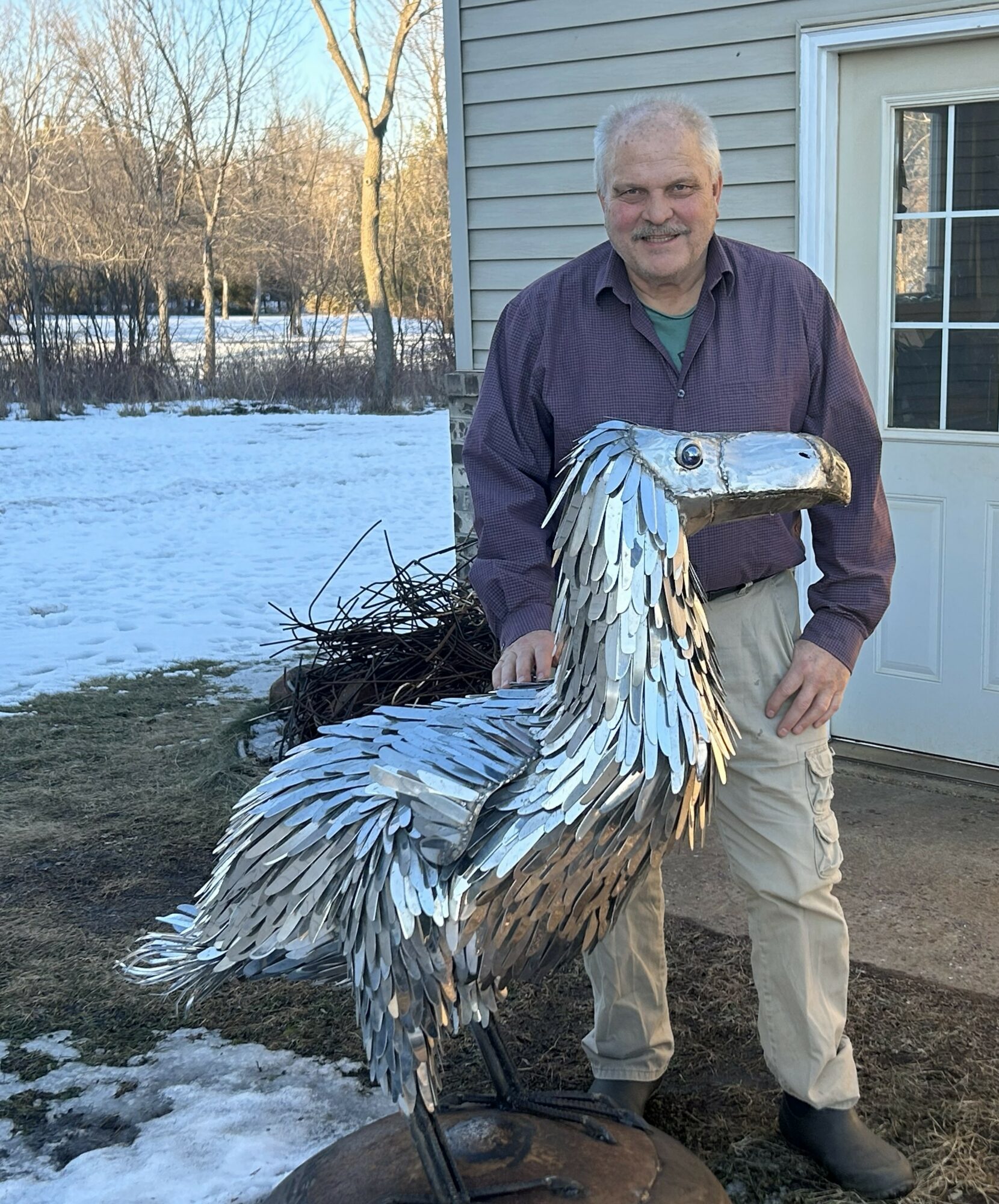 Man standing outdoors next to a large metal bird sculpture, with snow on the ground and a house in the background.
