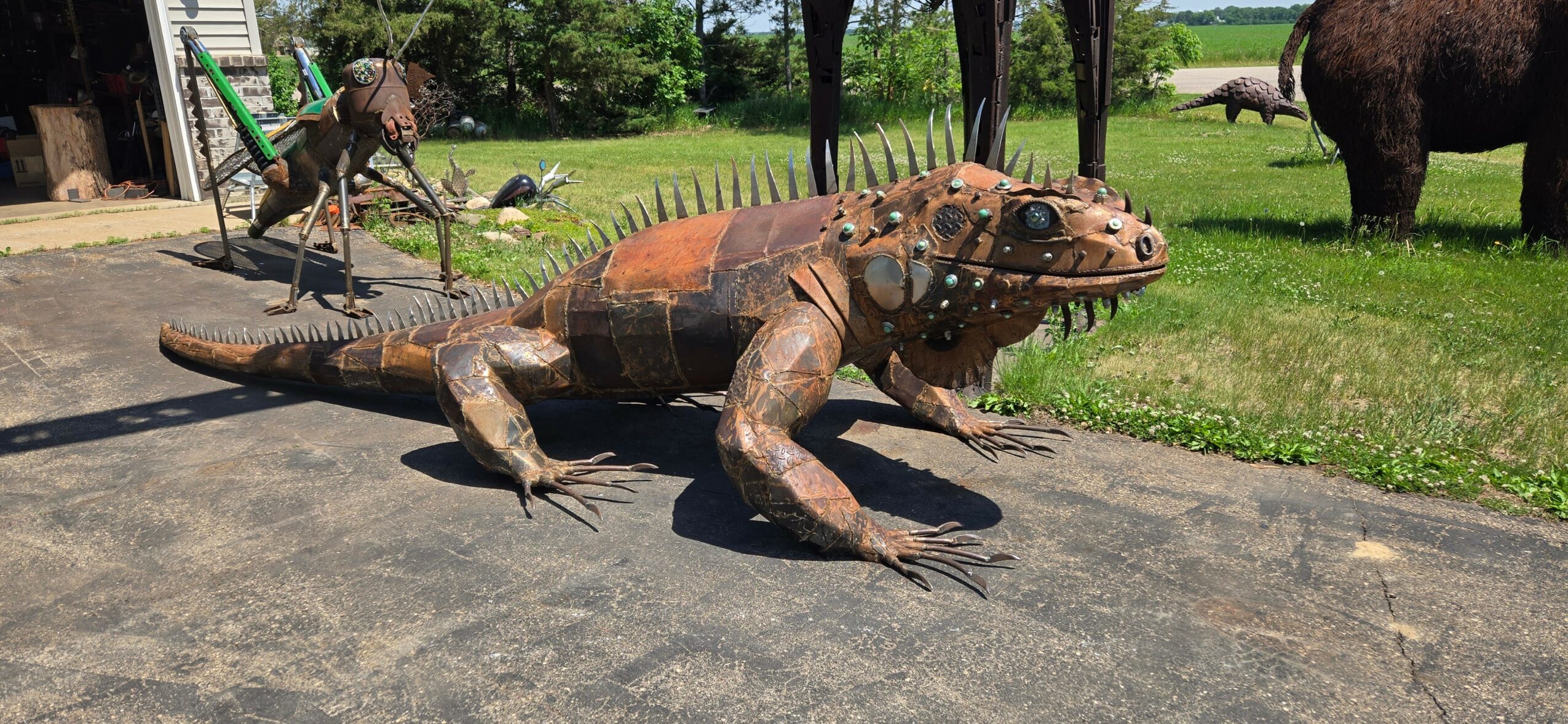 Large iguana sculpture on pavement with animals and trees in background.