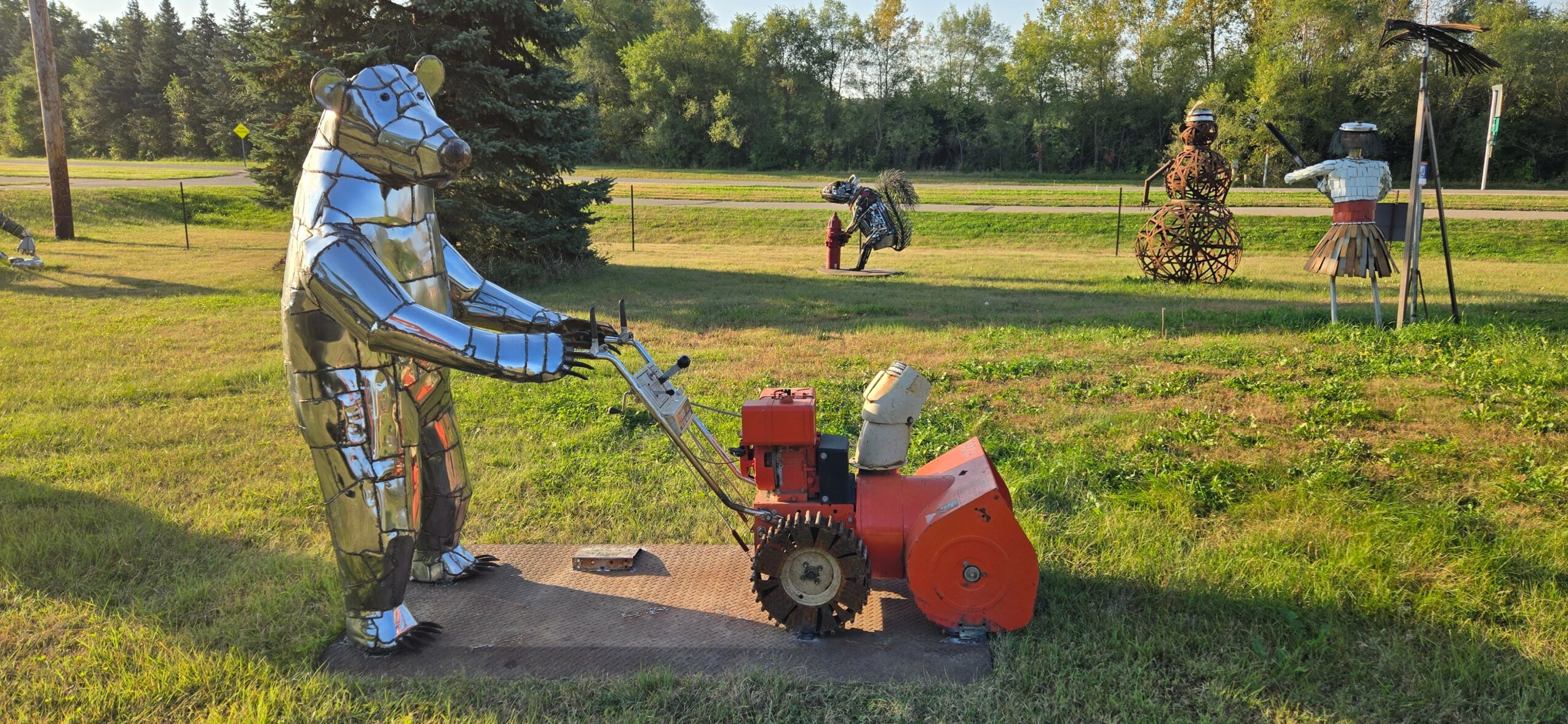 Person wearing protective gear operating a red and black machine outdoors on grass, with trees and people in the background.