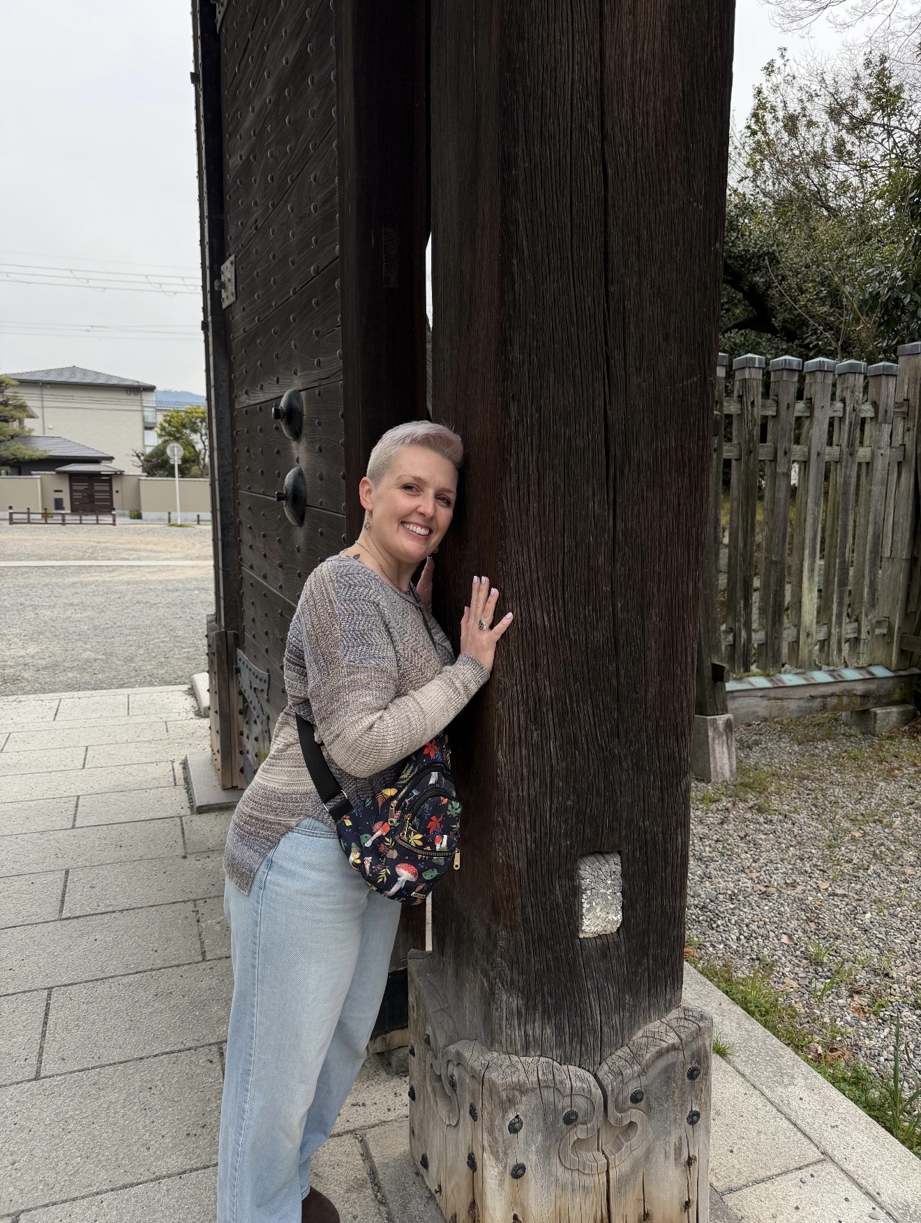 Woman leaning against a large wooden post outdoors, smiling, with a fence and trees in the background.