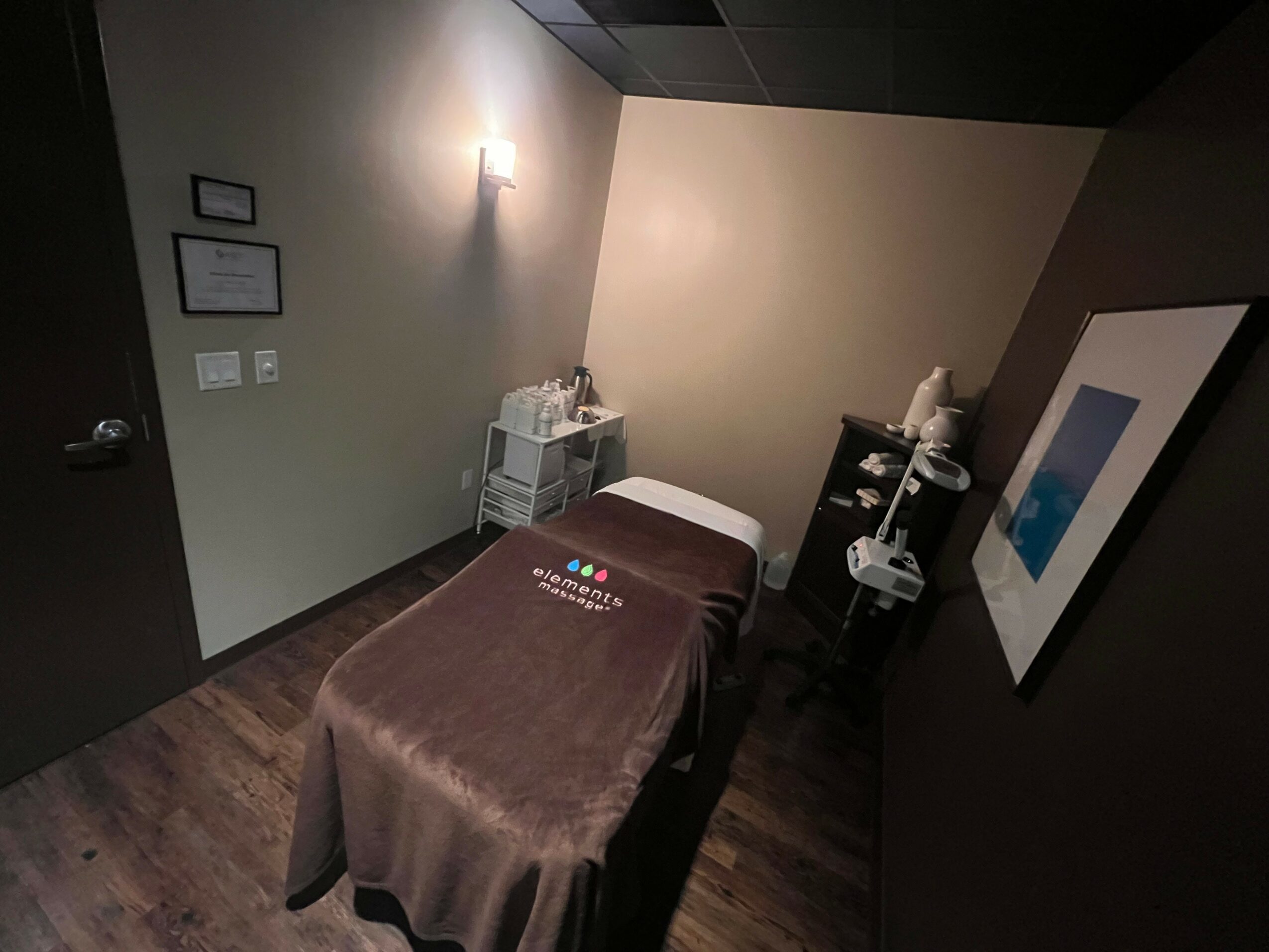Massage table covered with a brown sheet in a dimly lit room with artwork on the wall.