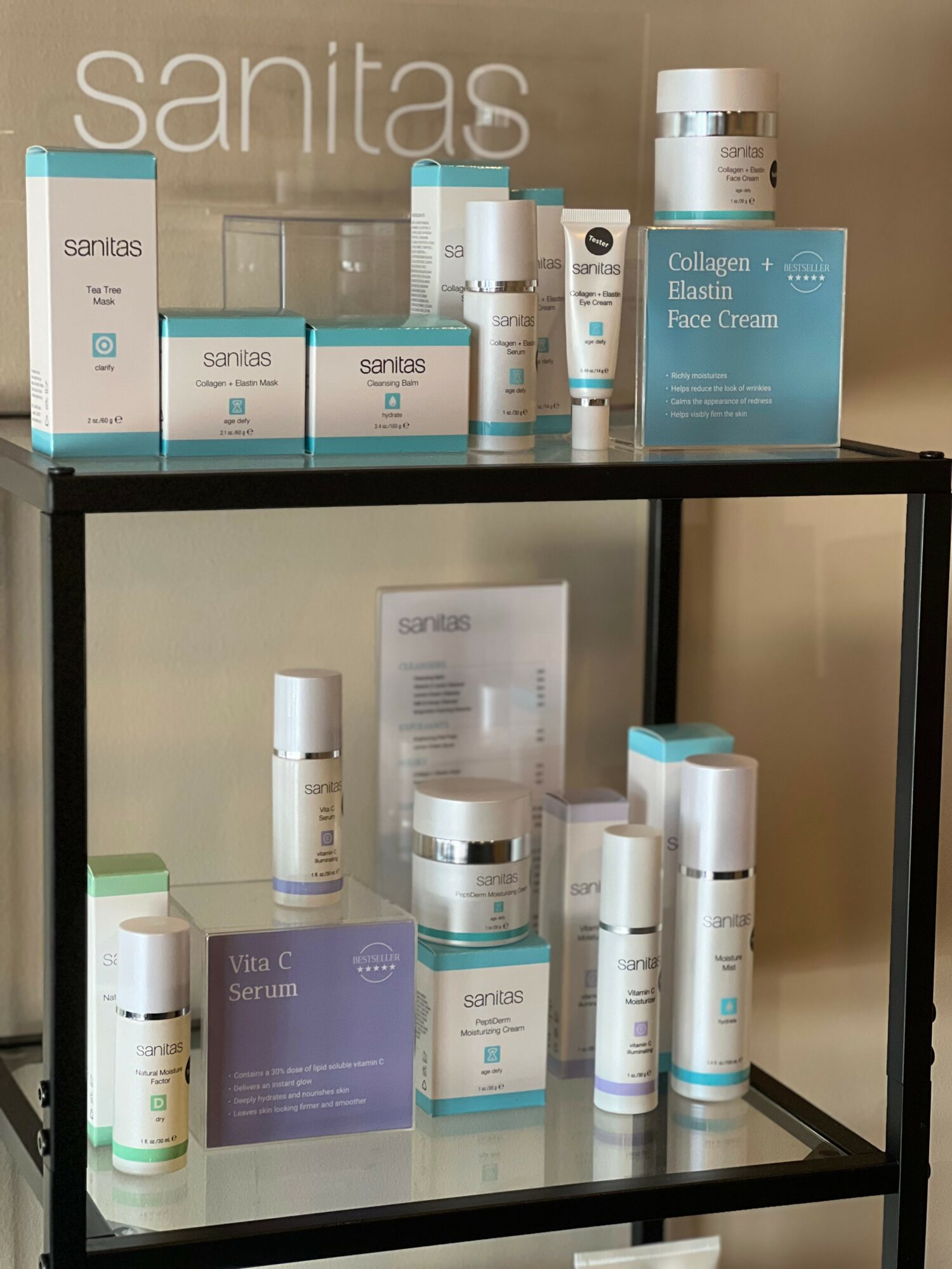 Shelf with skincare products, boxes, and bottles arranged on two levels, against a beige wall, with a 'sanitas' sign.