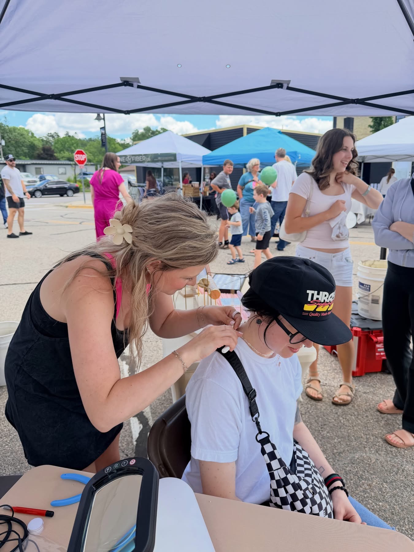 Woman helping a girl with glasses at an outdoor event under a canopy, with people and tents in background.