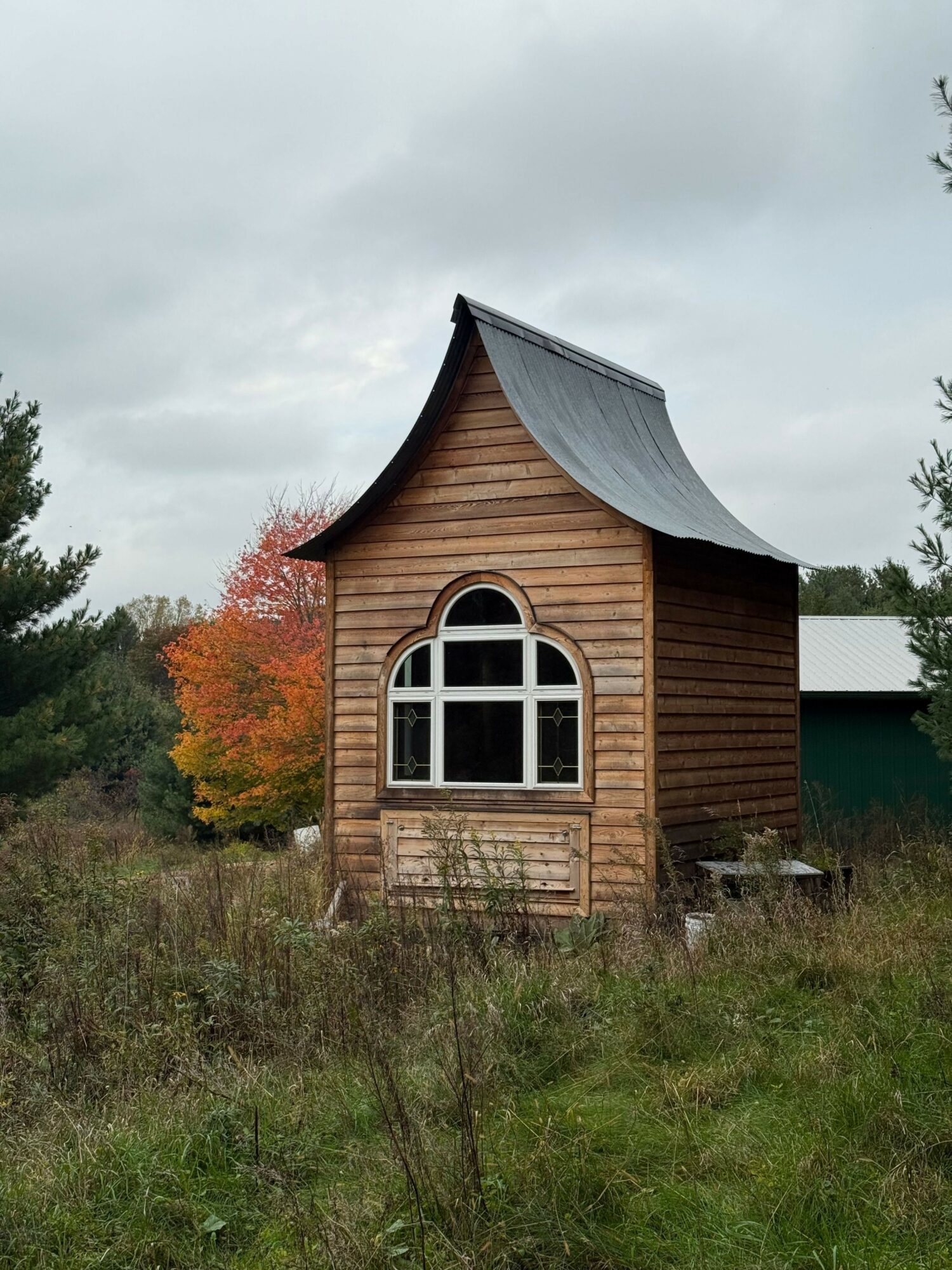 Small wooden house with a curved roof and arched window, surrounded by grass and trees, cloudy sky in background.