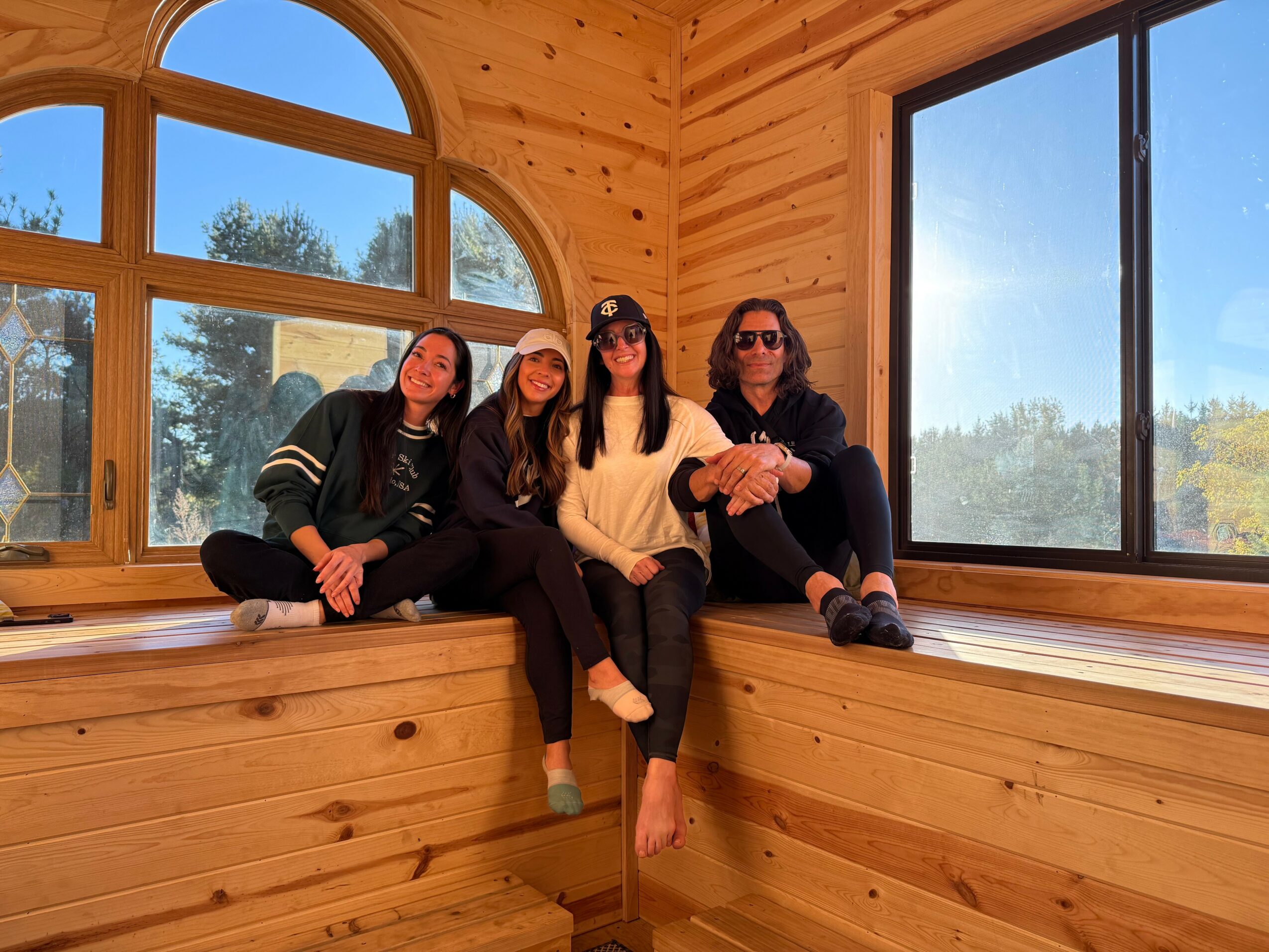Four women sitting on a wooden window seat inside a room with large windows and wooden walls, outdoors visible through windows.