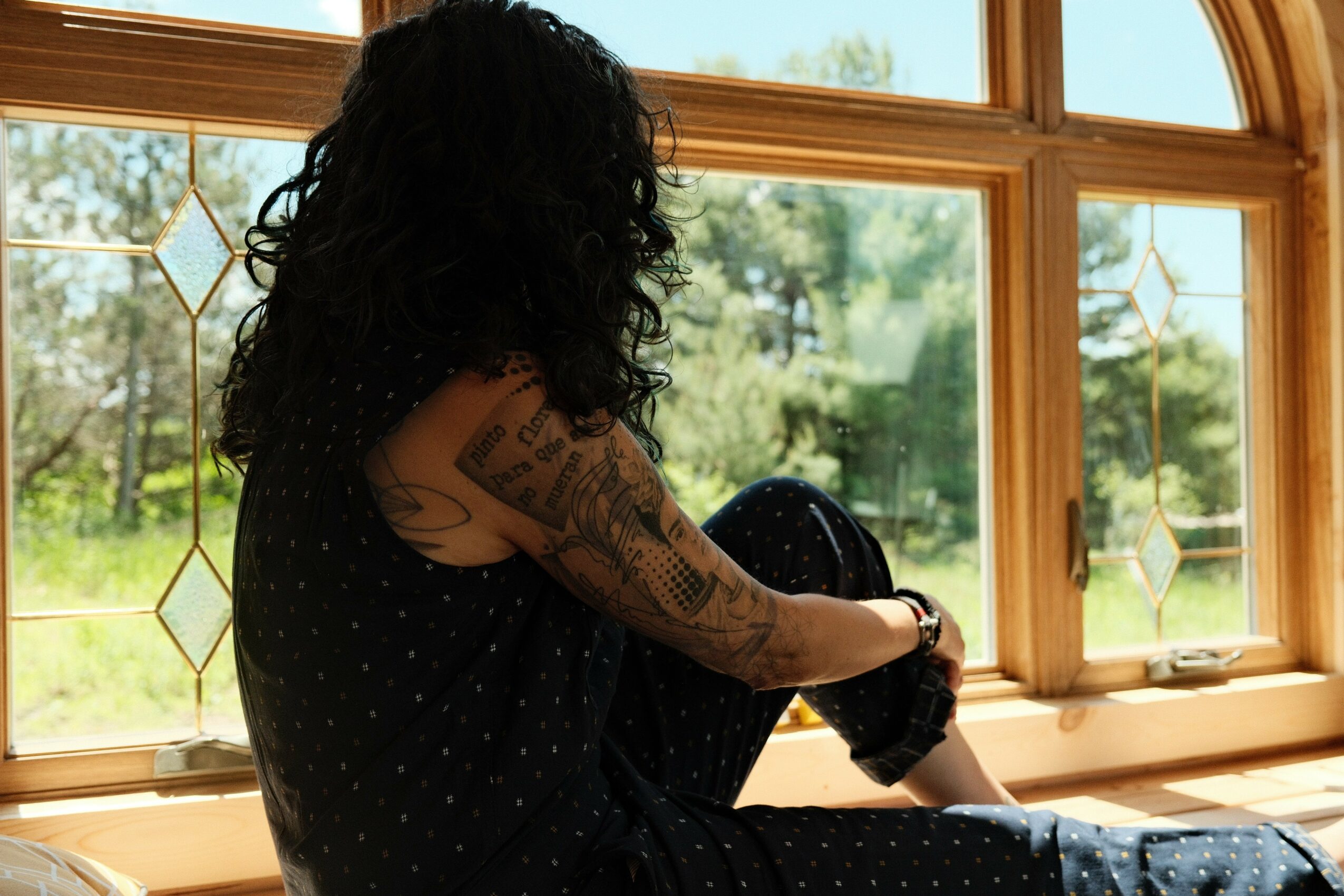 Person with curly hair and tattoos sitting by a window, looking outside at greenery.