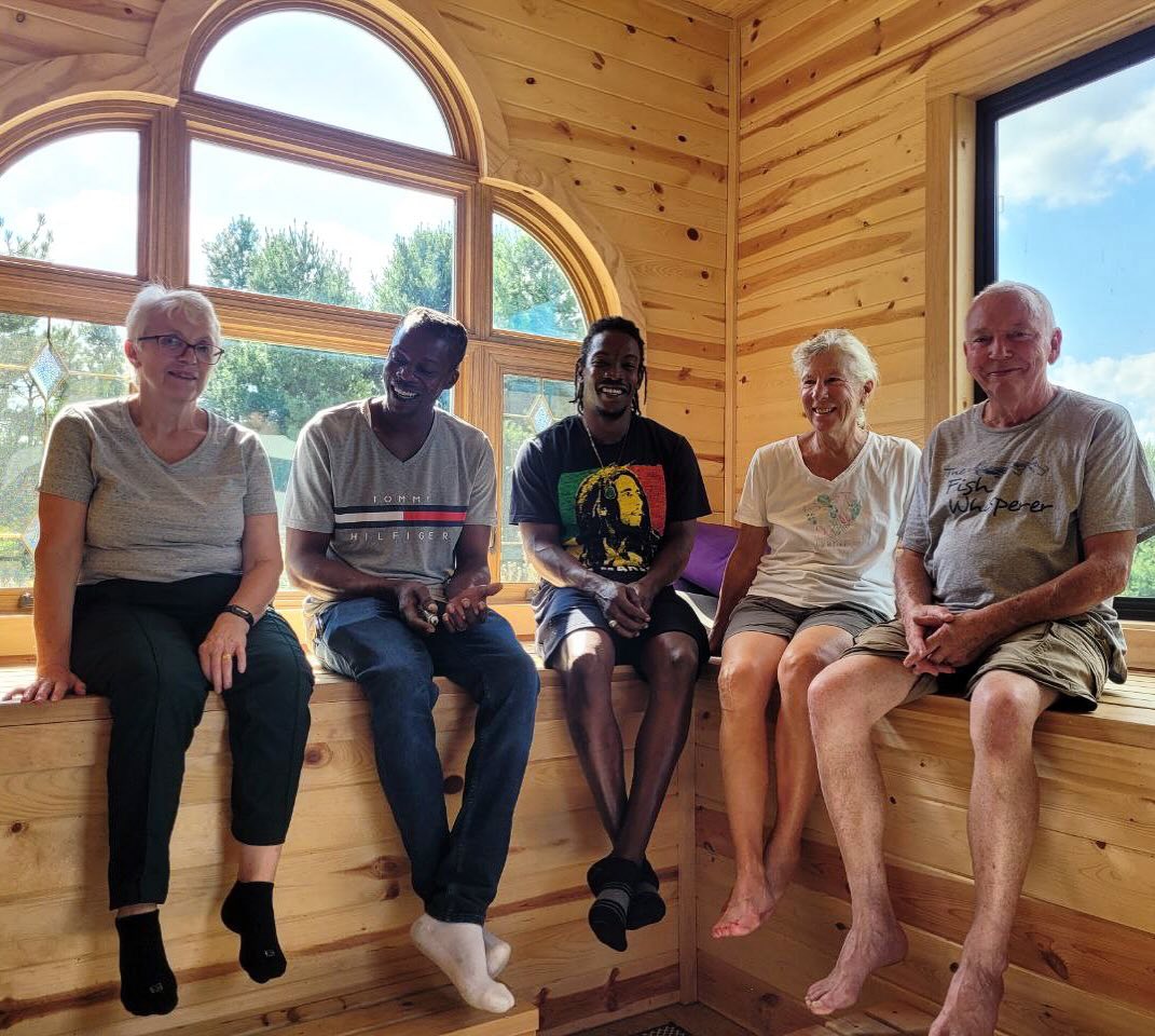 Five people sitting on a wooden window seat inside a room with large arched and rectangular windows, trees outside.
