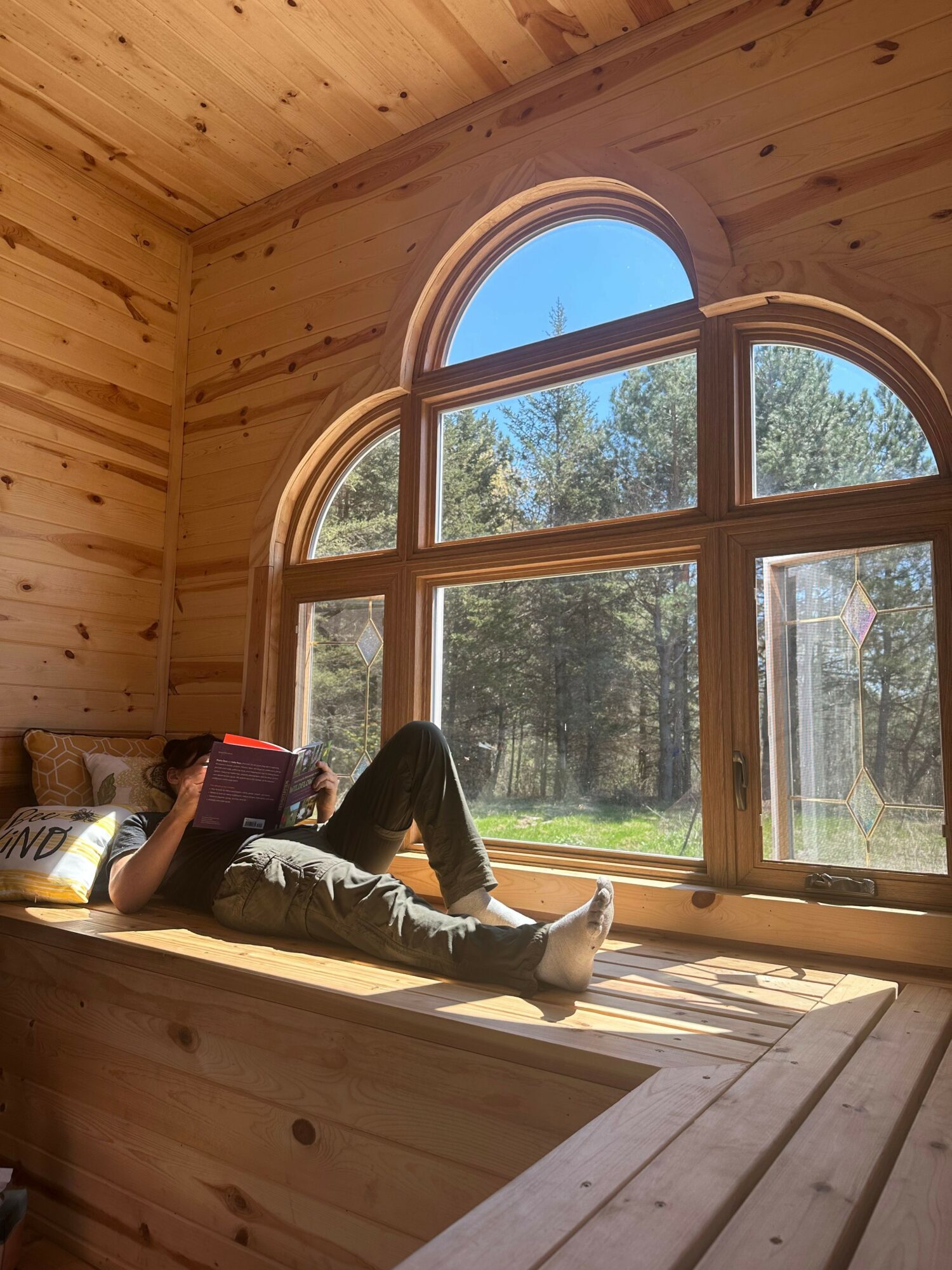 Person sitting on a windowsill inside a wooden room, with a large arched window showing trees and sky outside.