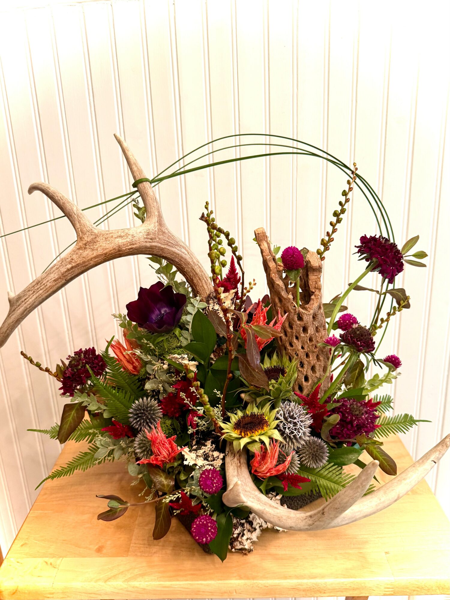 Arrangement of flowers and antlers with colorful blooms on a wooden surface, against a striped wall background.