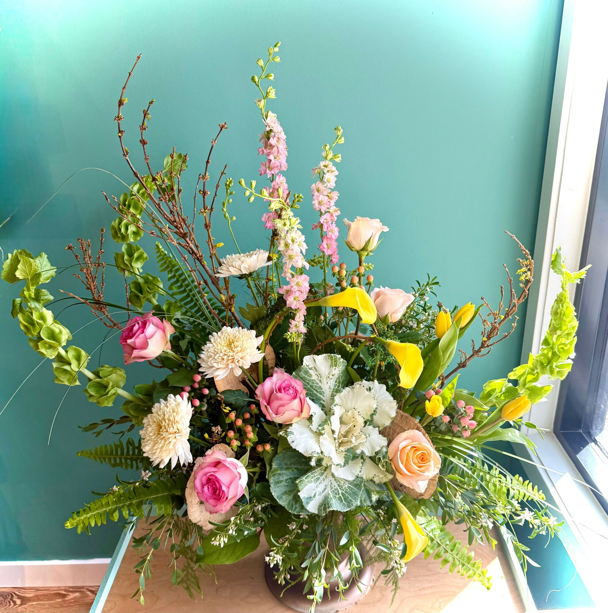 Flower arrangement with pink roses, yellow calla lilies, white flowers, and greenery in a glass vase near a window.
