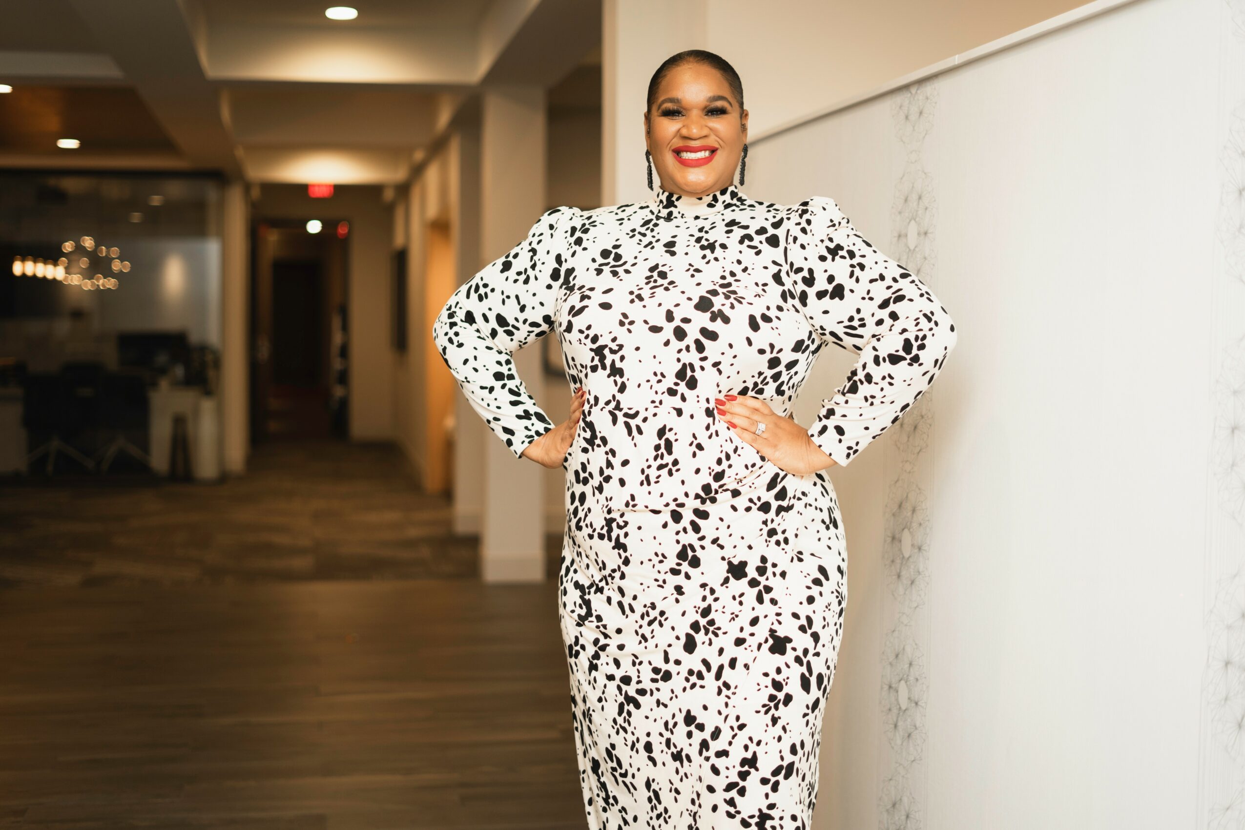 Woman in a black and white patterned dress standing with hands on hips, smiling, in an indoor setting.
